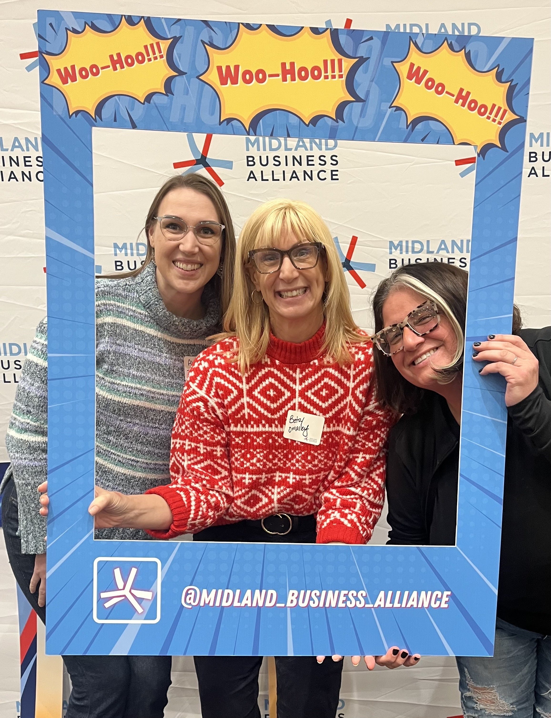 Three women smiling behind a large blue frame with comic-style yellow speech bubbles that say Woo-Hoo!!! and the social media handle @MIDLAND_BUSINESS_ALLIANCE, at an event for the Midland Business Alliance.