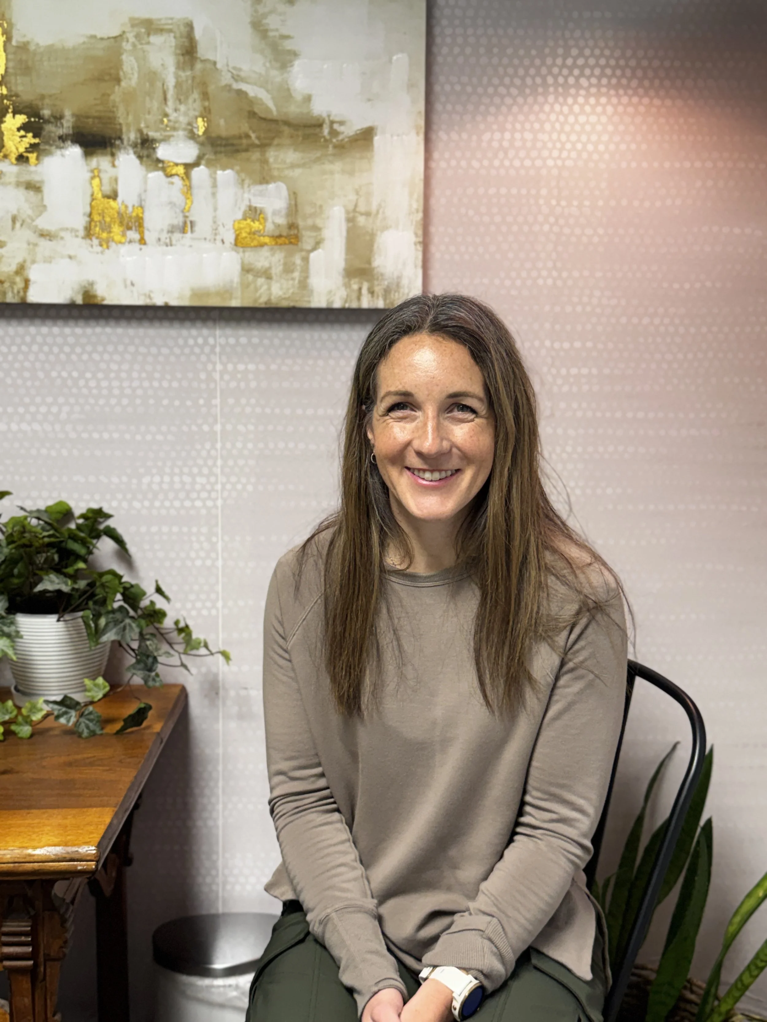 Pilates instructor Amber Rogers with long brown hair and freckles smiling while sitting at a table in Balanced Pilates and Wellness Boutique private room.