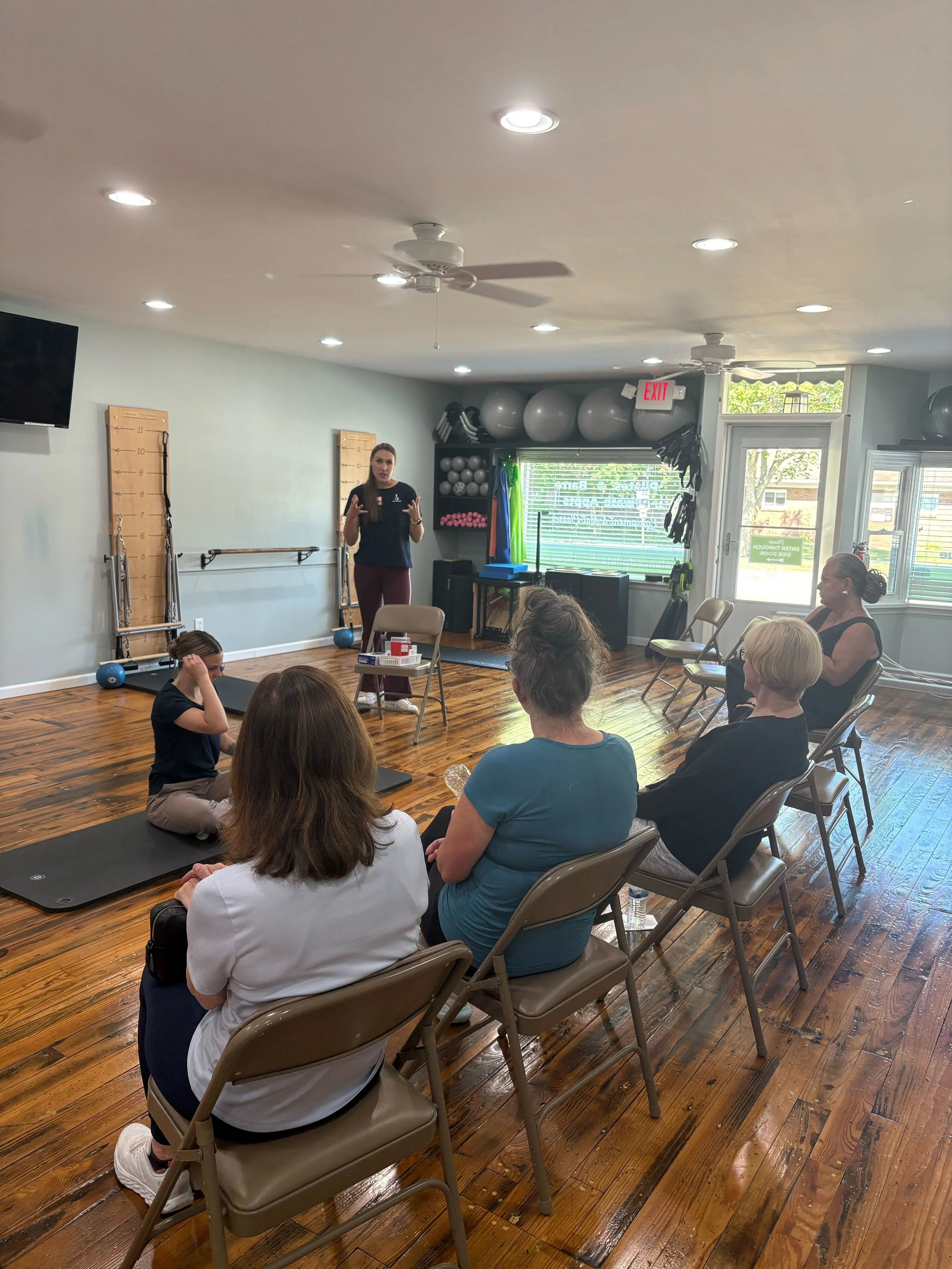 Indoor fitness or yoga class with women seated facing a woman instructor who is standing and talking, with fitness equipment and balls on the wall, and sunlight coming through large windows.