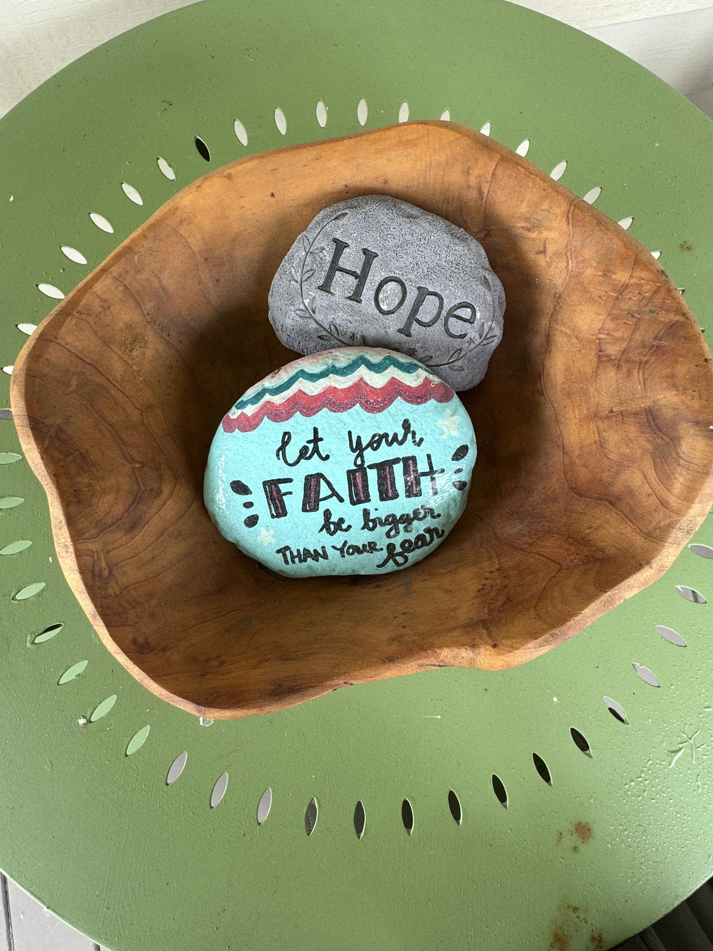 Decorative painted stone with the word 'Hope' and a painted inspirational message on a wooden bowl, placed on a green perforated metal table.