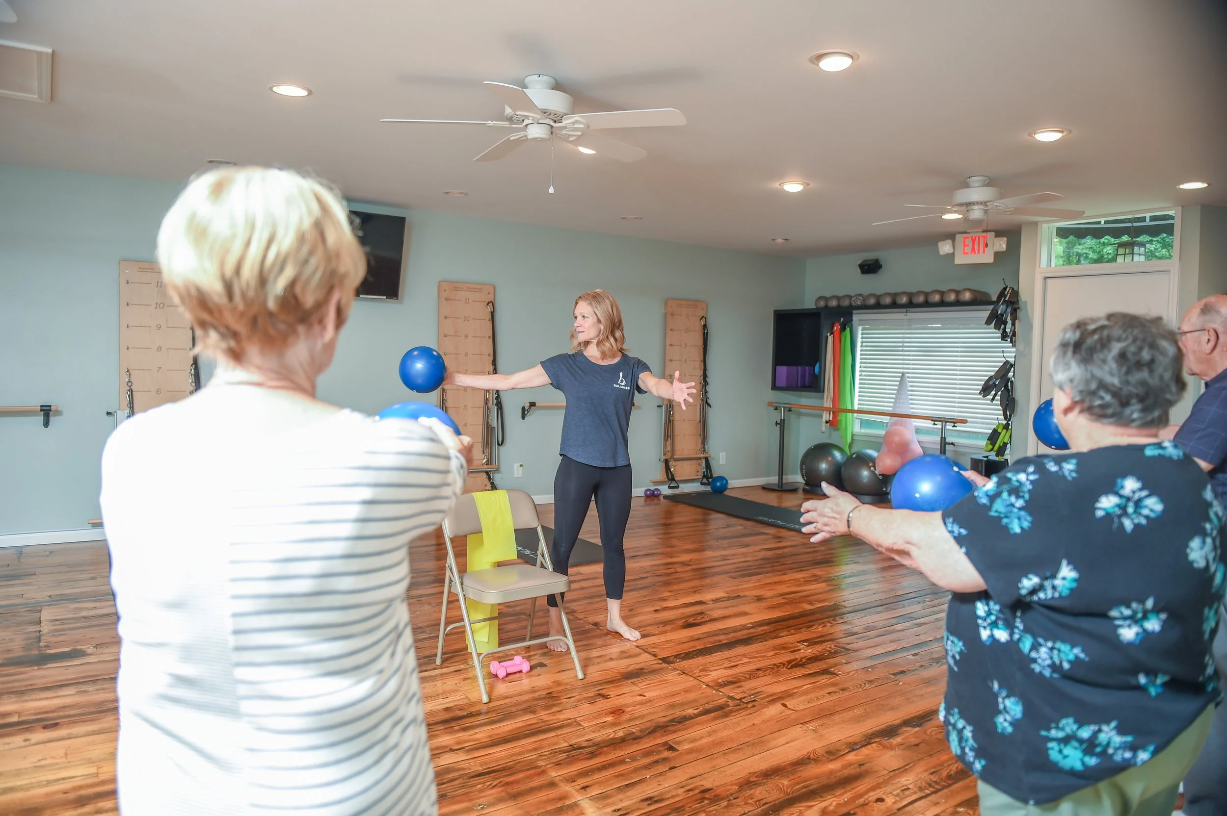 A woman leading a group exercise class for seniors in a Pilates studio in Midland, Michigan, demonstrating with a blue exercise ball while the seniors follow along, holding smaller balls