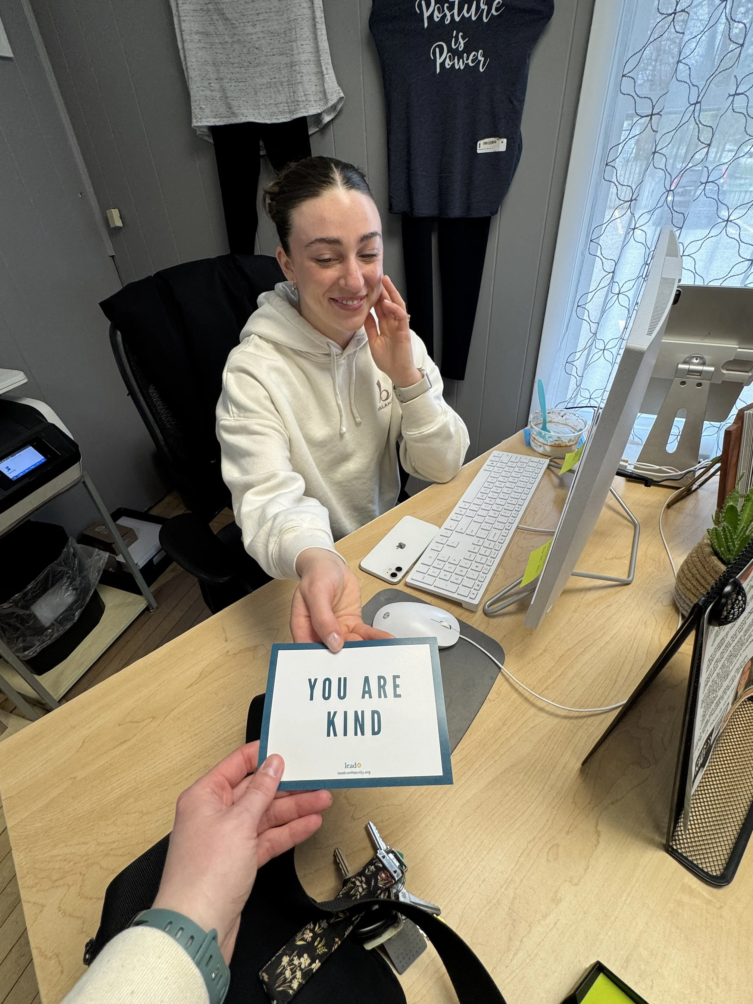 A woman sitting at a desk smiling, with a card that says "You Are Kind" being held by a person in the foreground. The desk has a computer, a smartphone, and a few decorative items.