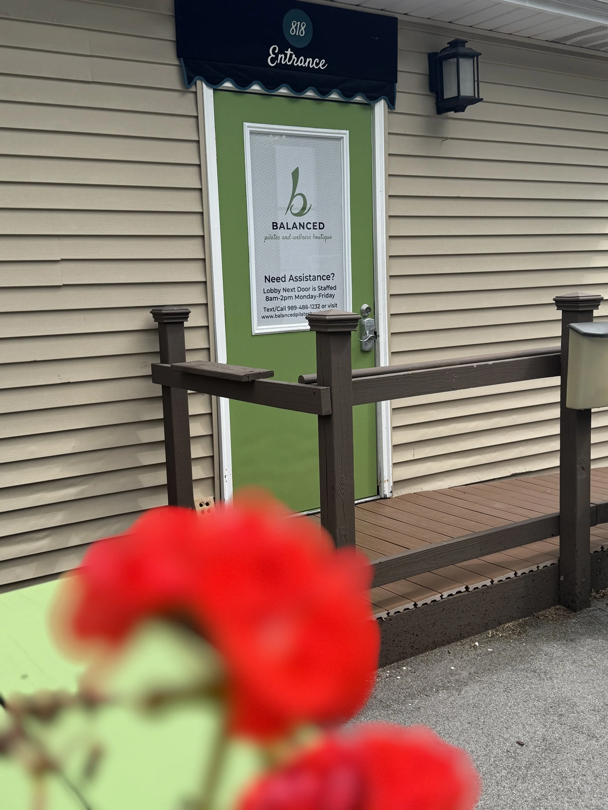 A green door with white trim and a sign that reads 'Balanced Pilates and Wellness Boutique' with contact info, at an entrance to a building. The door has a keypad lock and is framed by beige siding. There is a black wall lantern next to it, and a woo