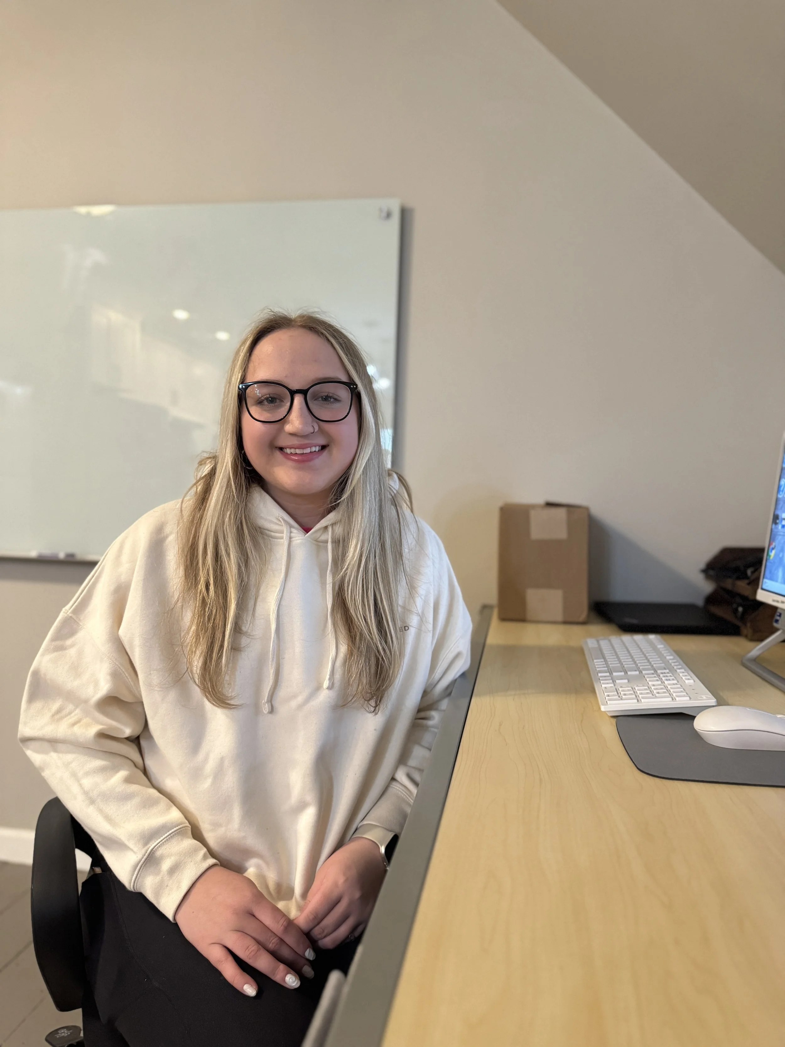 A young woman with long blonde hair, glasses, and a nose ring, smiling and sitting at a desk with a keyboard, mouse, and computer monitor in an office setting.