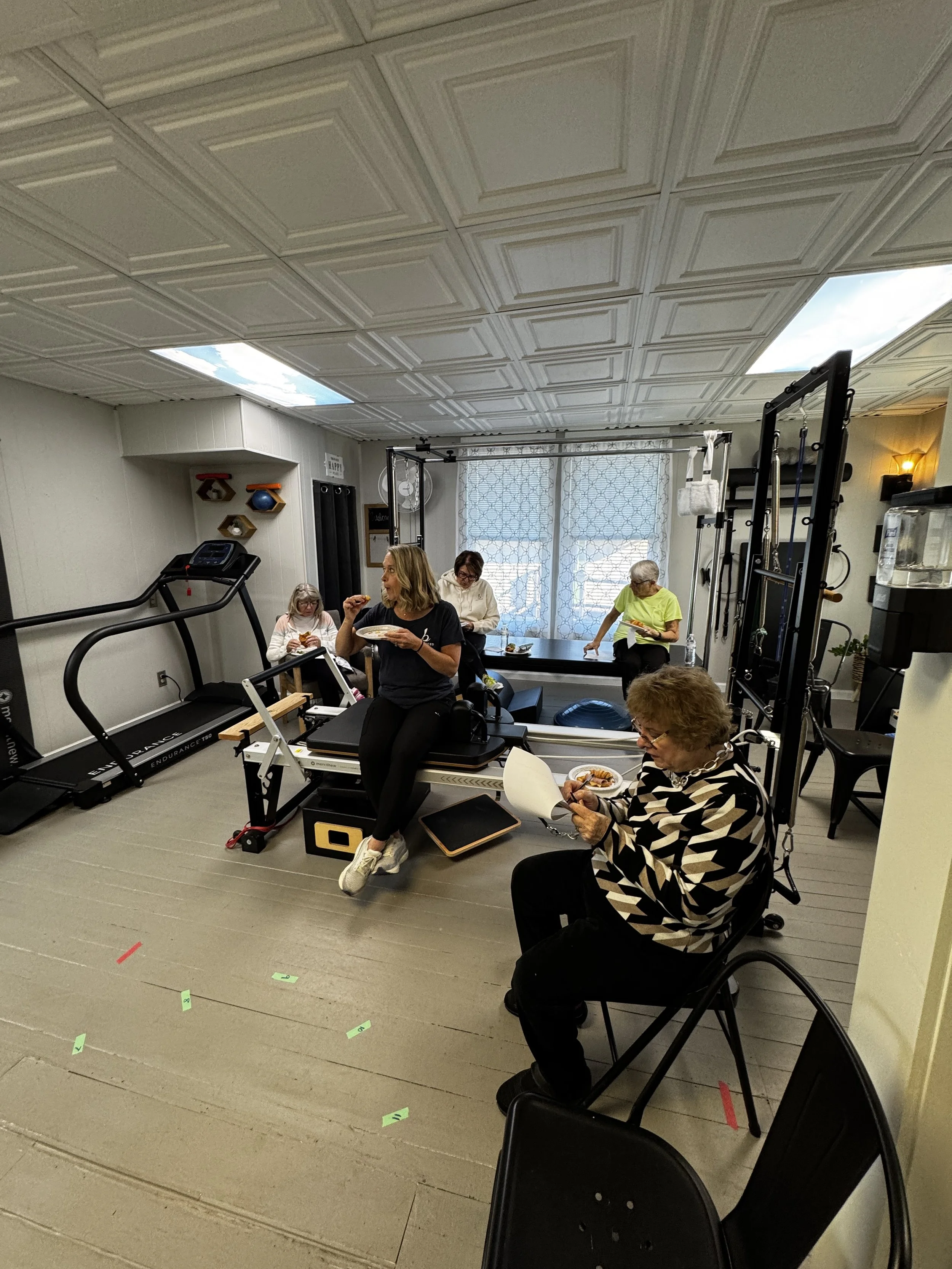 Multiple people in a fitness studio, some sitting and eating, others using exercise equipment. The room has a ceiling with patterned tiles and windows with sheer curtains. There are workout machines, weights, and a water bottle visible.