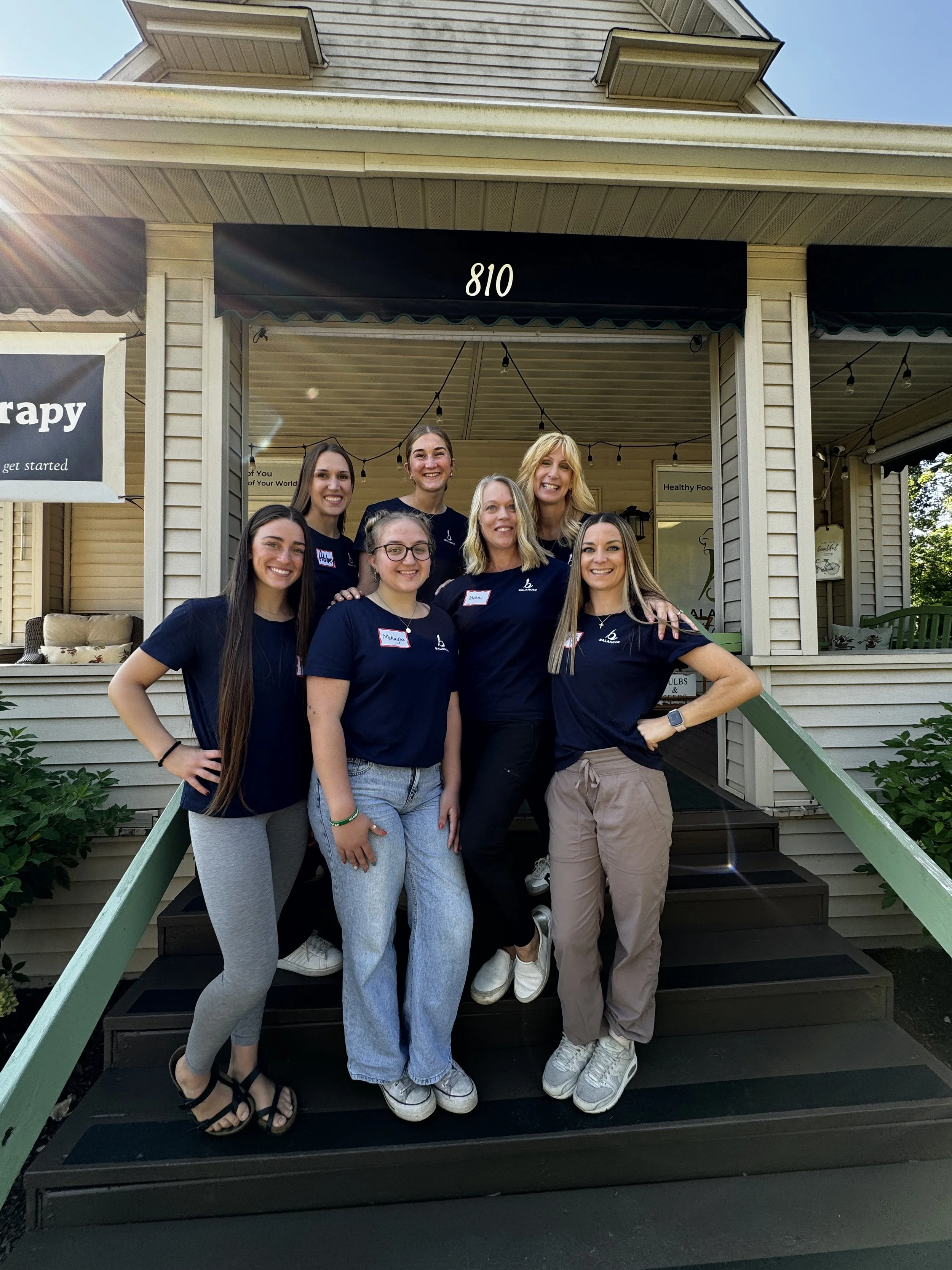 Group of seven women standing on steps outside a building with black awning and house number 810, smiling for a photo.