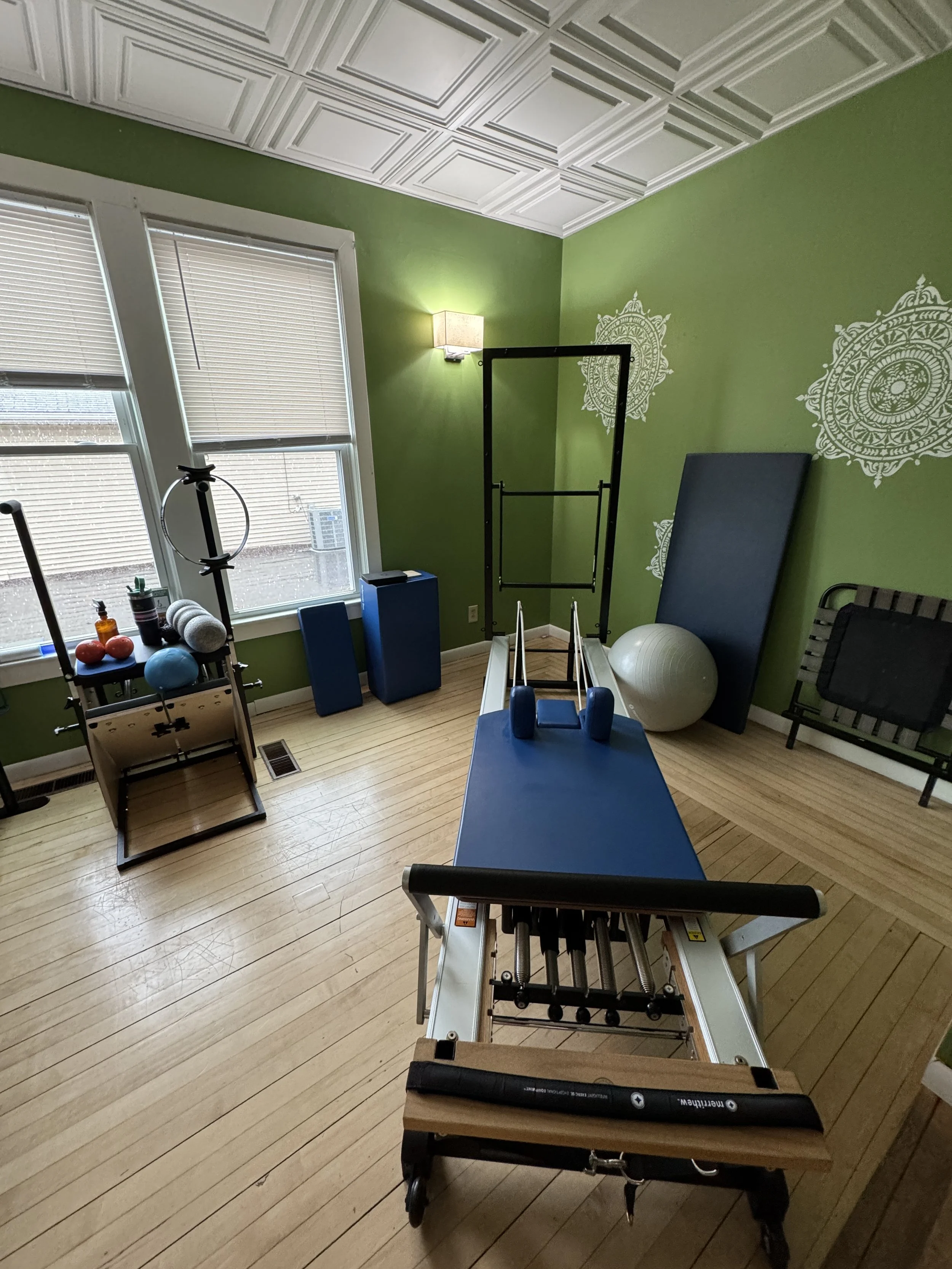 Room with green walls and a decorative ceiling, containing fitness equipment such as a rowing machine, exercise ball, mat, and a treadmill, with large windows and a wall-mounted light at Balanced Pilates and Wellness Boutique in Midland, Michigan.