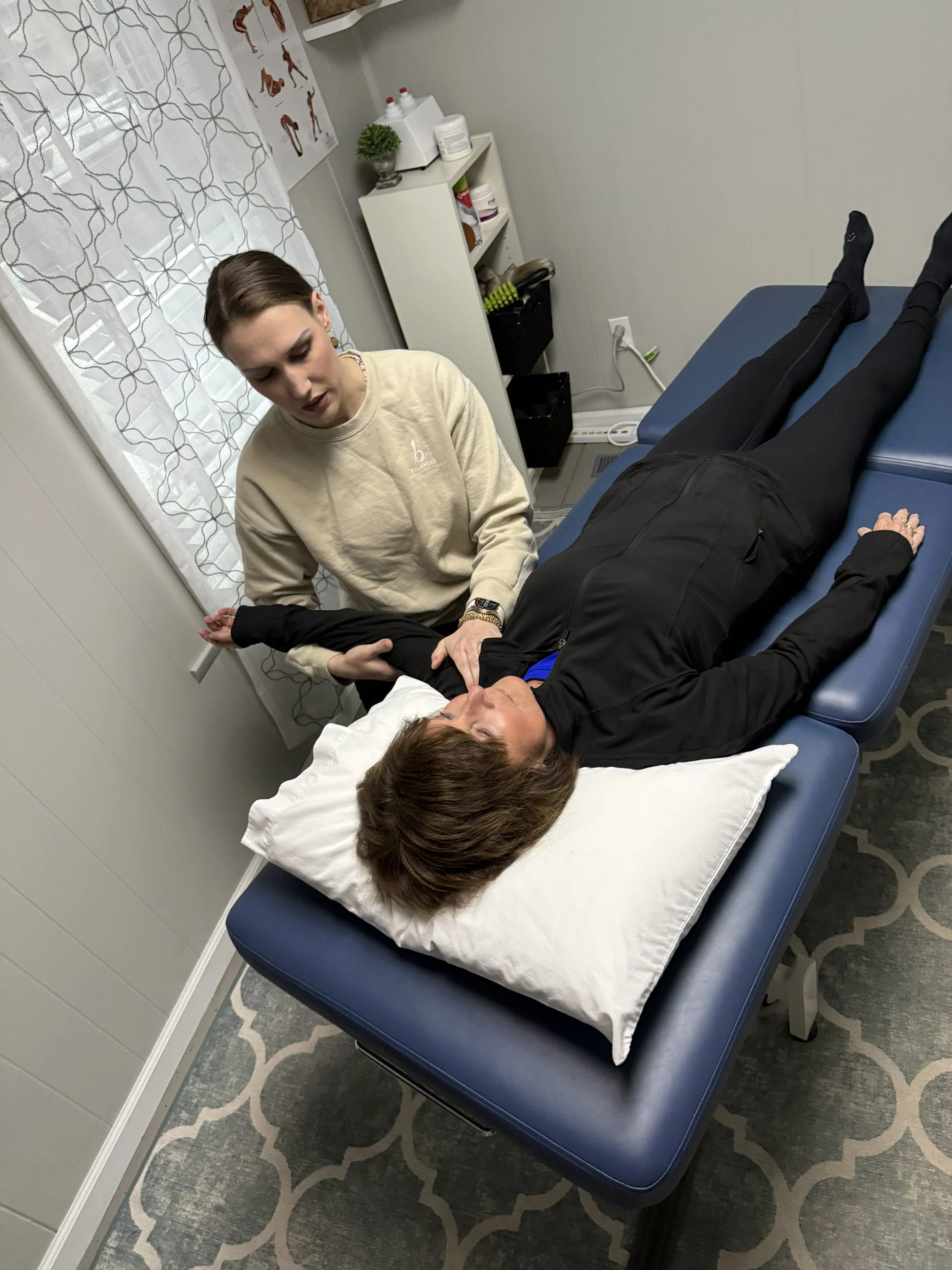 Physical therapist is working with a patient. The room has gray walls, a patterned gray and white rug, a small white shelf with medical supplies, and a window with sheer curtains decorated with a geometric pattern.