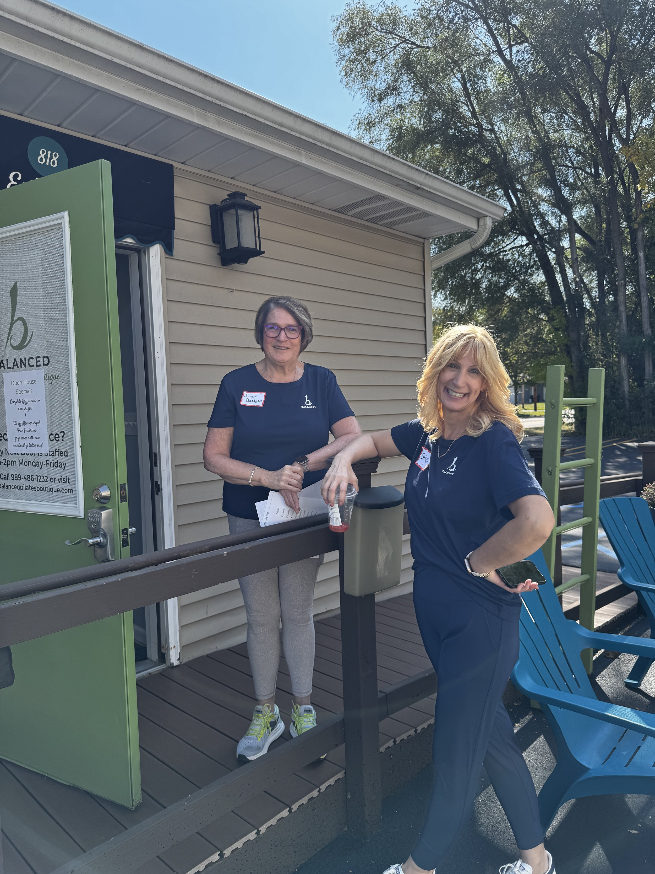 Two women standing outside a beige building near a green vending machine and blue chairs. One woman with short gray hair and glasses, and another with long blonde hair, both smiling.