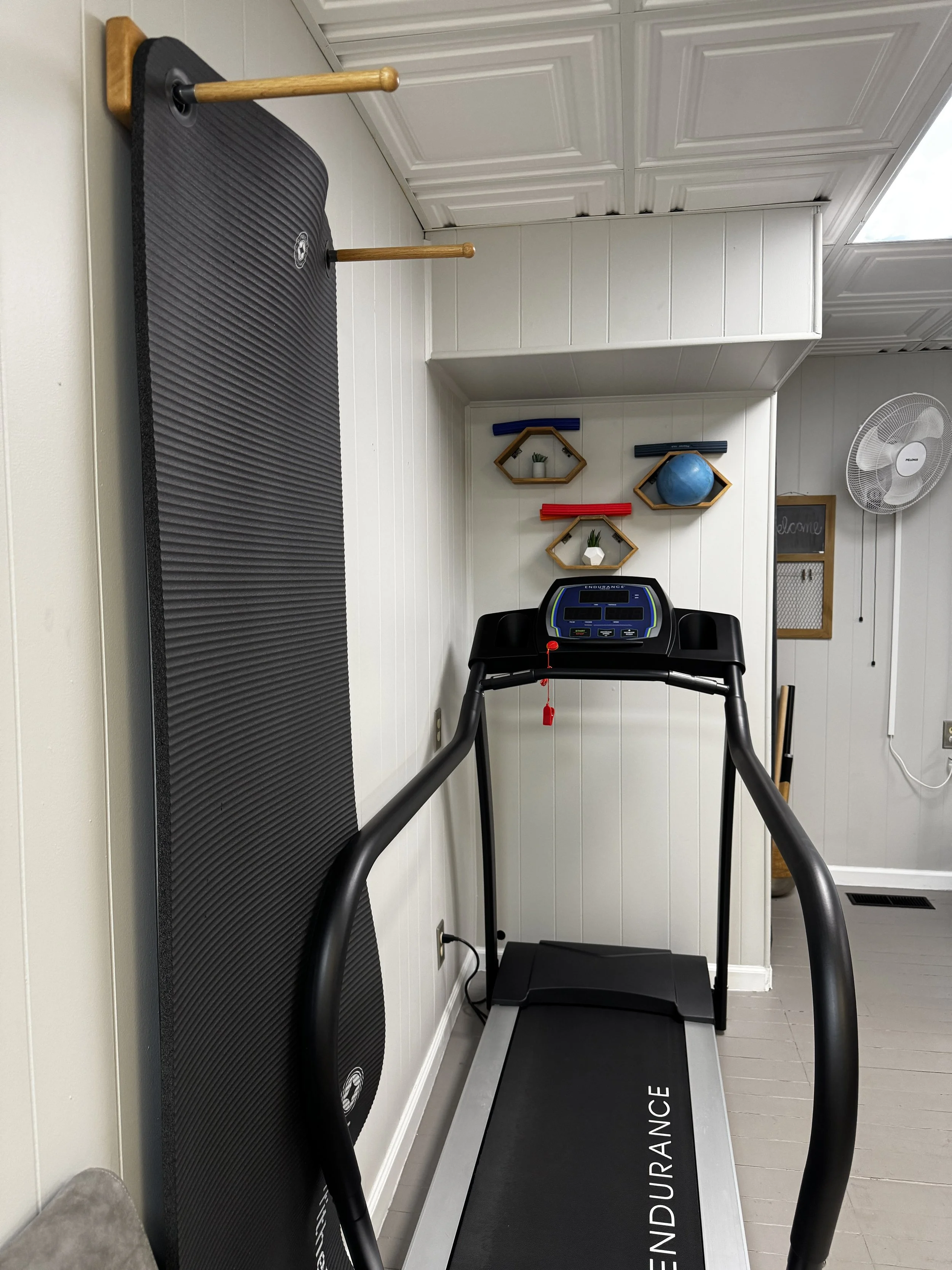 A treadmill in a private physical therapy gym with black handles and a display panel, placed indoors near white paneled walls and a fan on the wall, with a black exercise mat labeled 'ENDURANCE' on the floor.