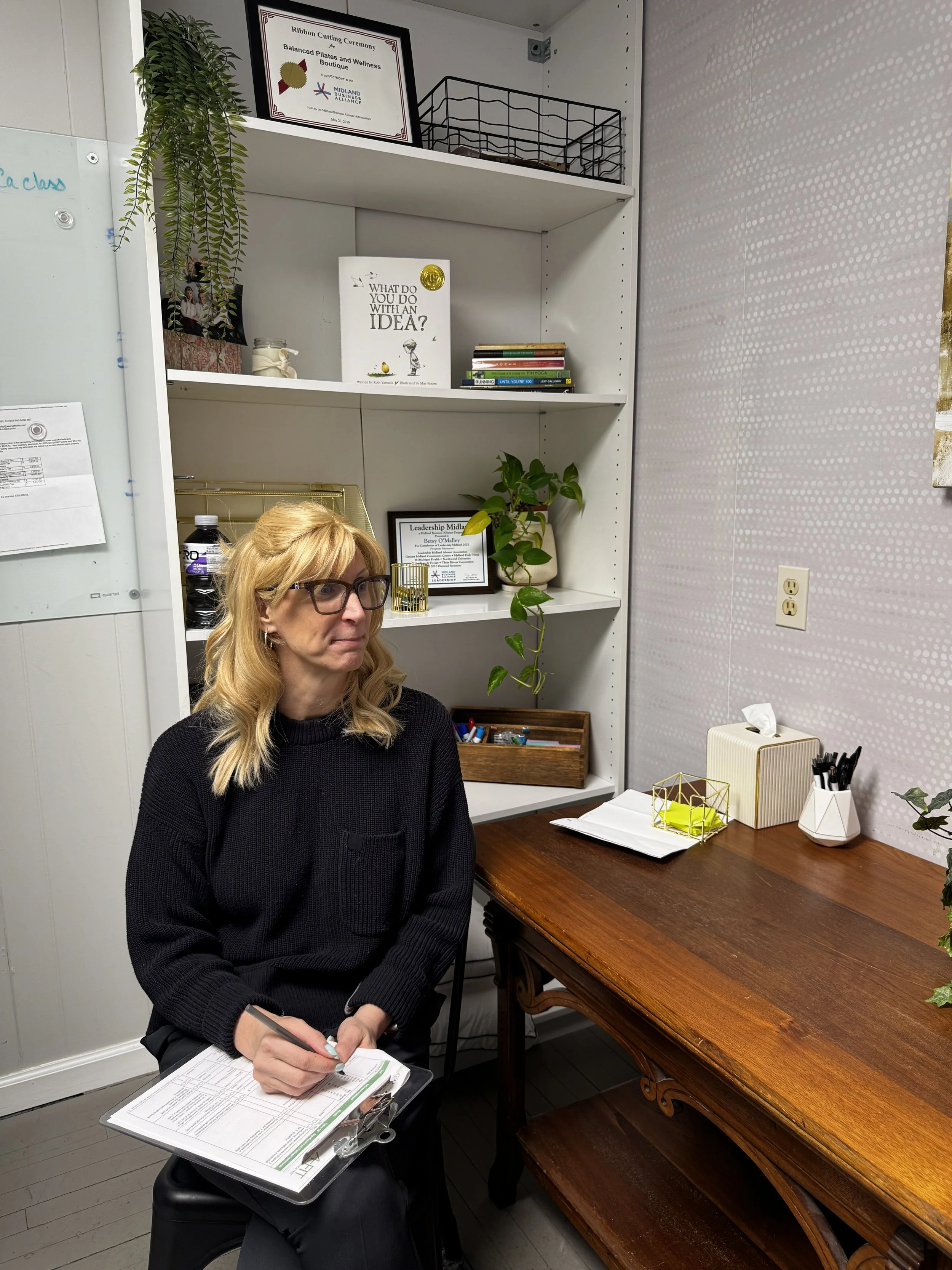 A woman with blonde hair and glasses sitting at a polished wooden desk with papers and a clipboard, holding a pen. Behind her are white shelves with decorative items, books, and green plants, next to a gray wall with an electrical outlet.