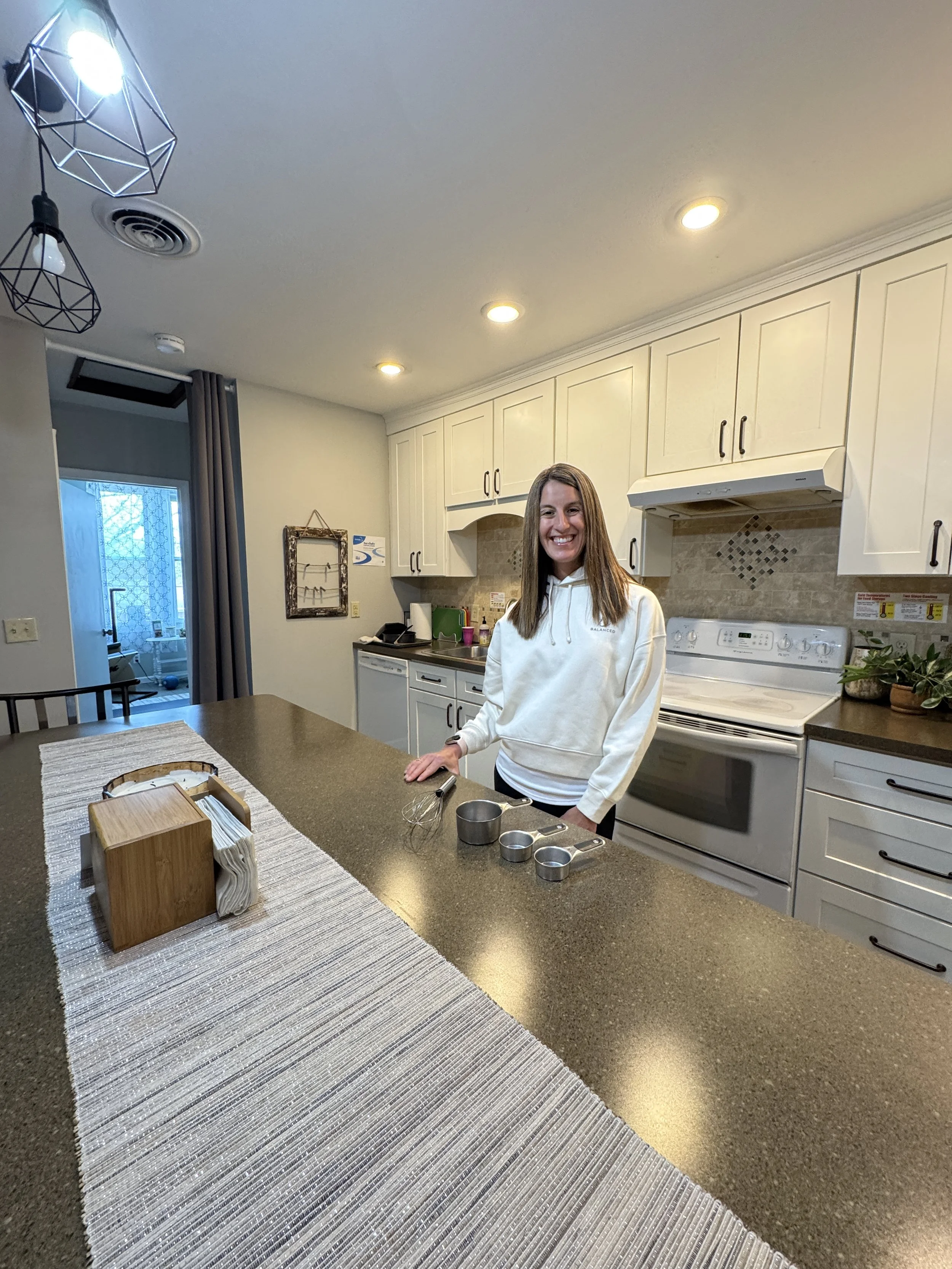A smiling woman in a white hoodie stands in Balanced kitchen with white cabinets and beige backsplash. There are three small metal measuring cups on the counter, a wooden tissue box holder, and a striped gray rug on the brown countertop.