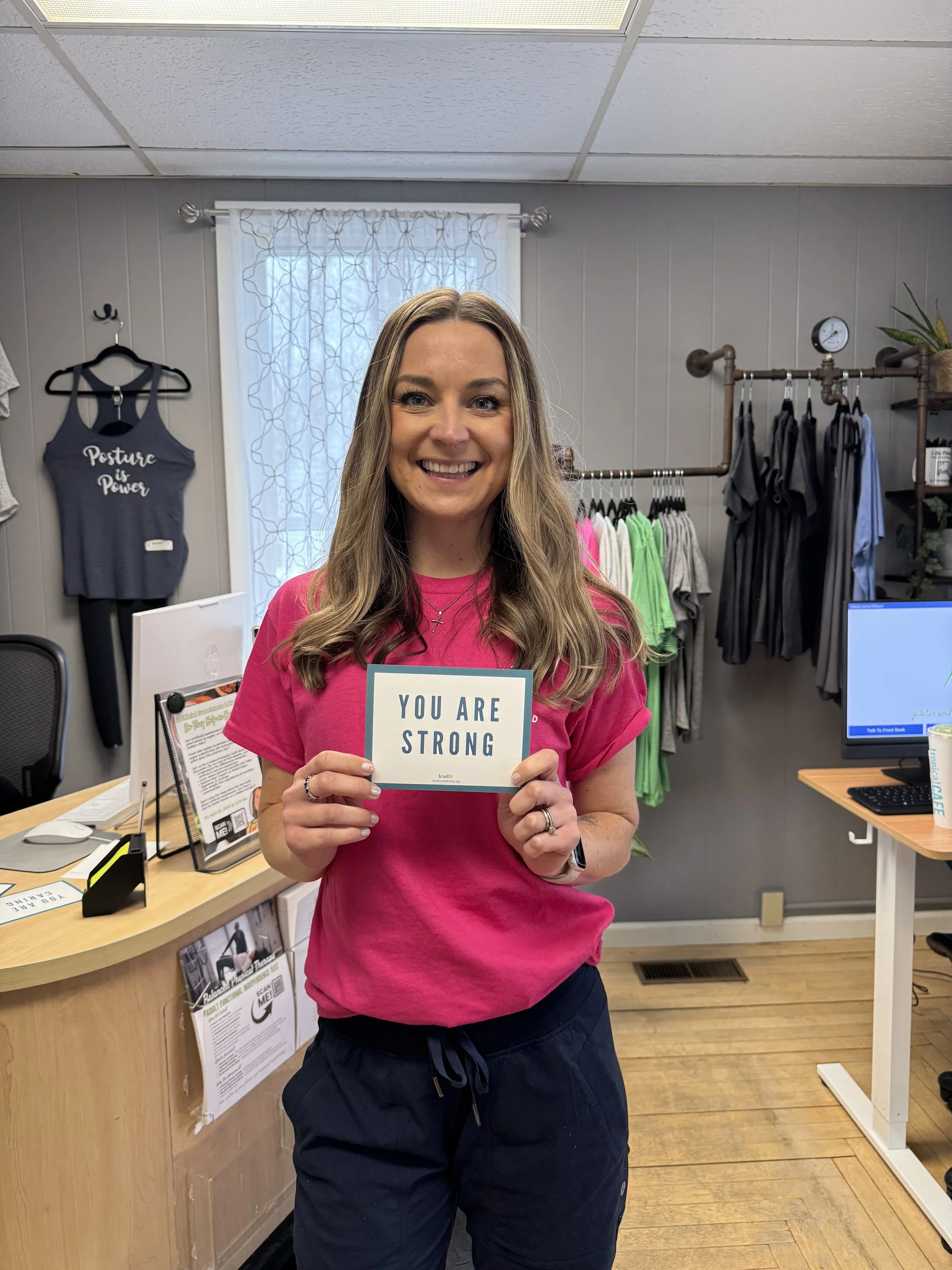 A woman wearing a pink T-shirt holding a sign that says 'You Are Strong' inside Balanced Pilates and Wellness Boutique in Midland, Michigan. There are clothing racks and a desk with various items and posters in the background.