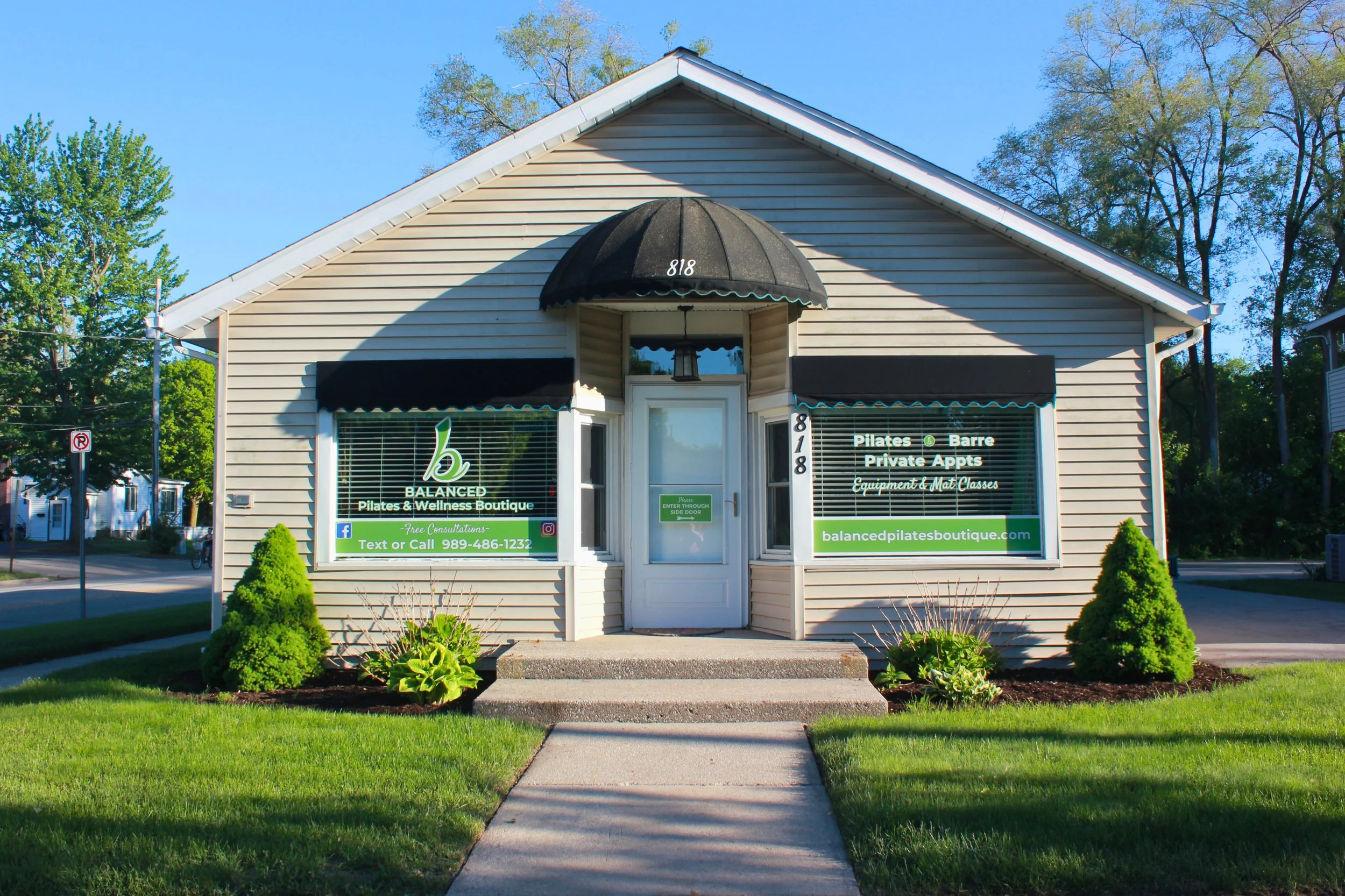 Front of a small beige building with a sign for a Pilates and wellness boutique with the number 818, and a green lawn with small bushes and a sidewalk leading to the door at Balanced Pilates and Wellness Boutique in Midland.