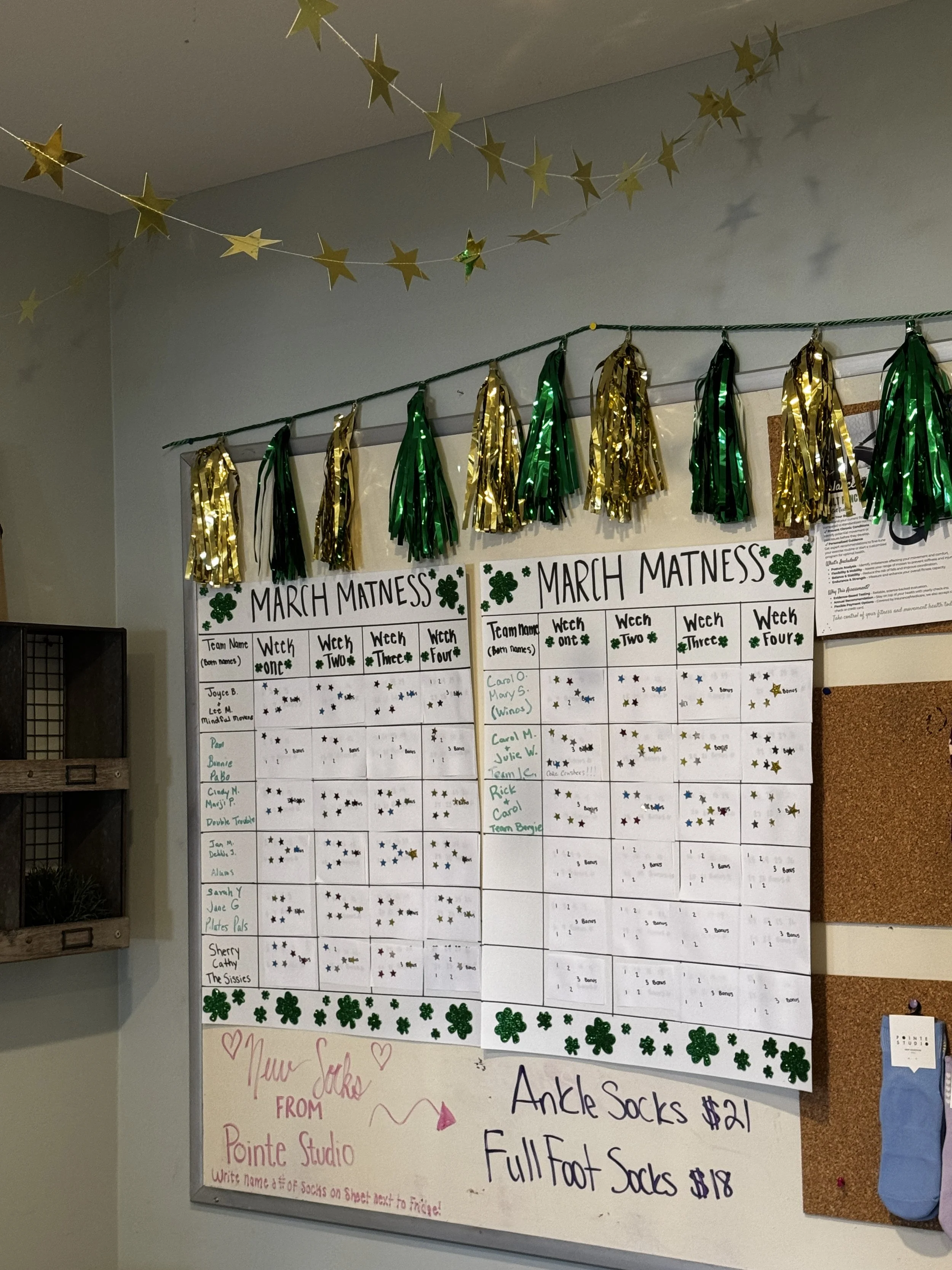 Whiteboard with handwritten March Matness Pilates Challenge schedule decorated with green and gold shamrock and star stickers, and a note about ankle socks and full foot socks for sale, next to a corkboard and a blue sock.