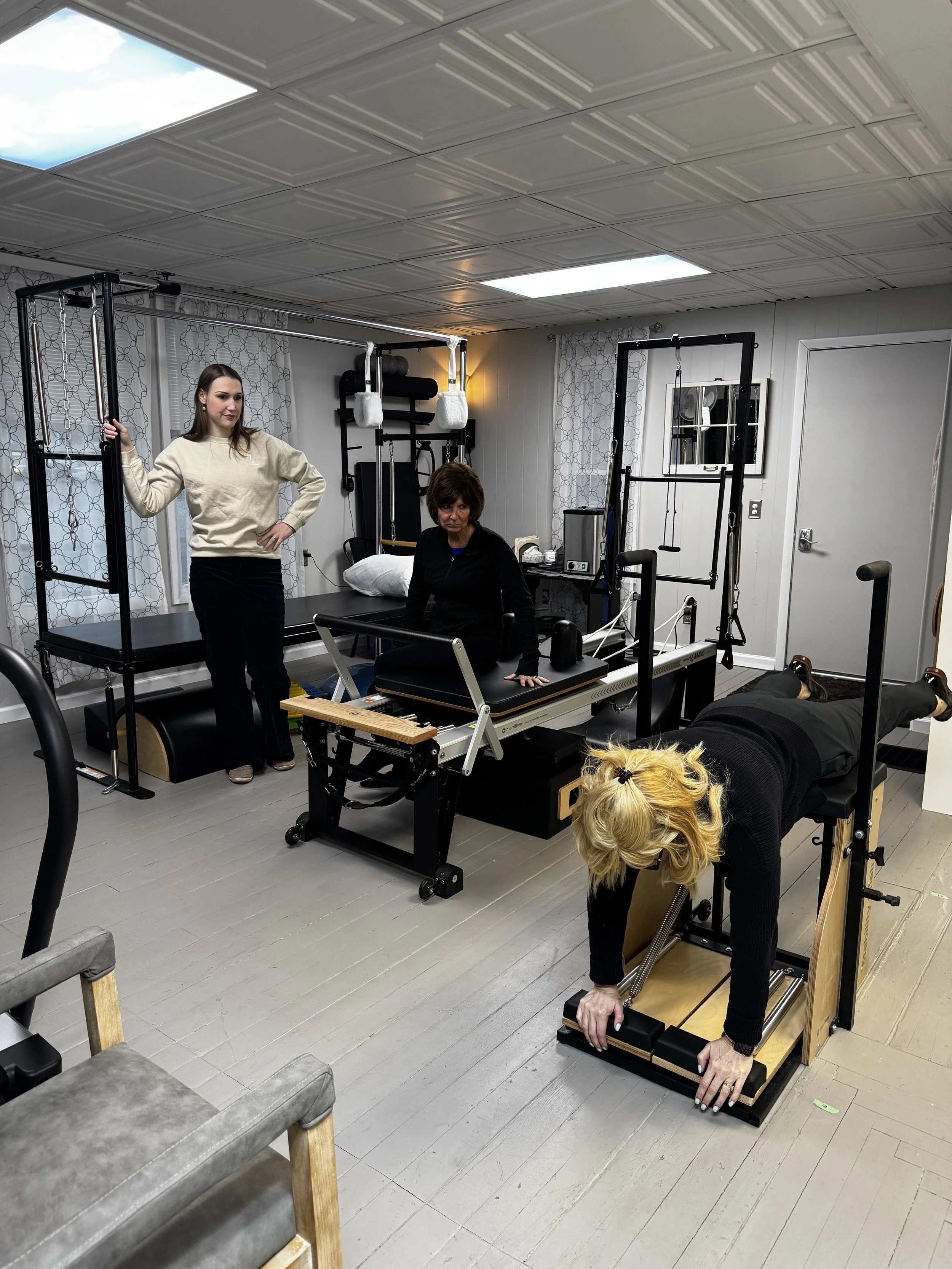 A physical therapy session in a clinic with two women using Pilates equipment. One woman is lying sideways on a reformer machine, holding onto the bars, while the other woman, standing nearby, is observing. The room has white walls, patterned curtain