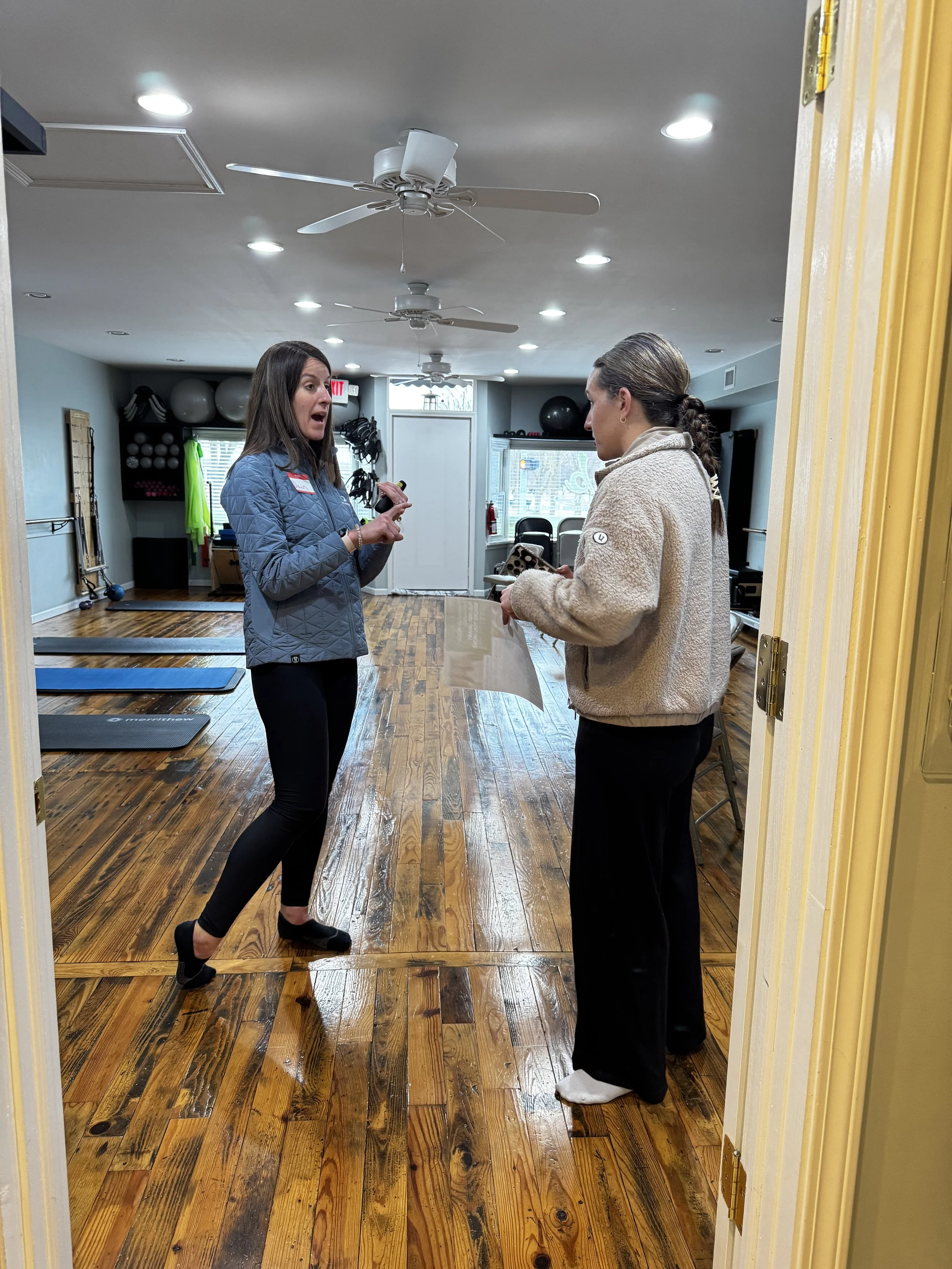 A woman in a blue jacket is talking to another woman in a cream-colored fleece jacket inside a Midland Pilates studio.