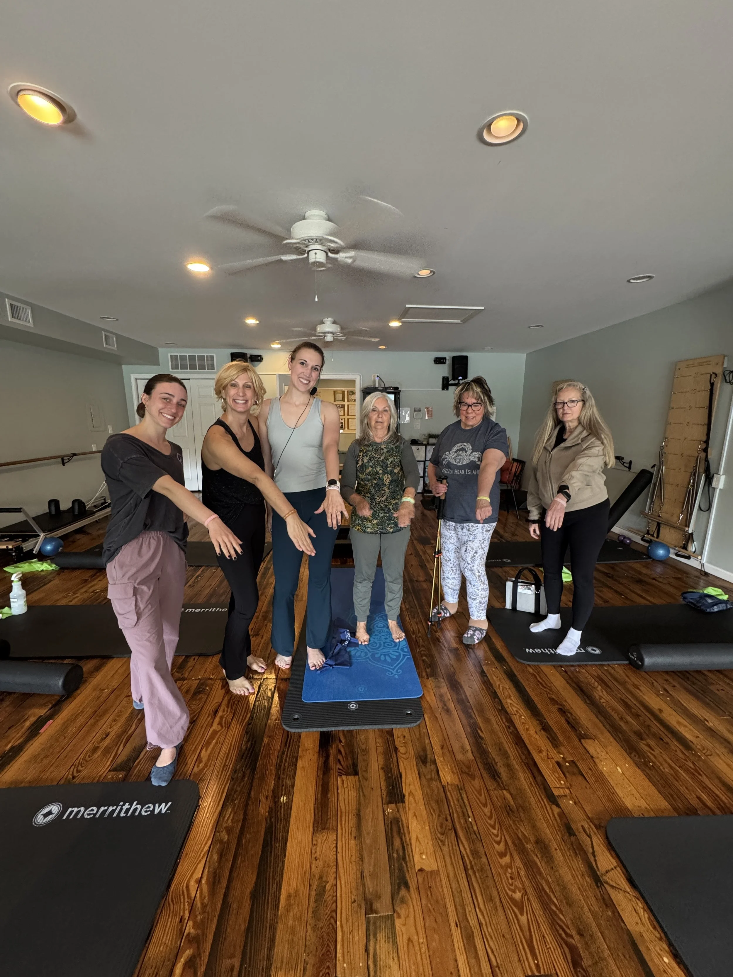 Group of six women standing on yoga mats in a fitness studio, smiling at the camera.