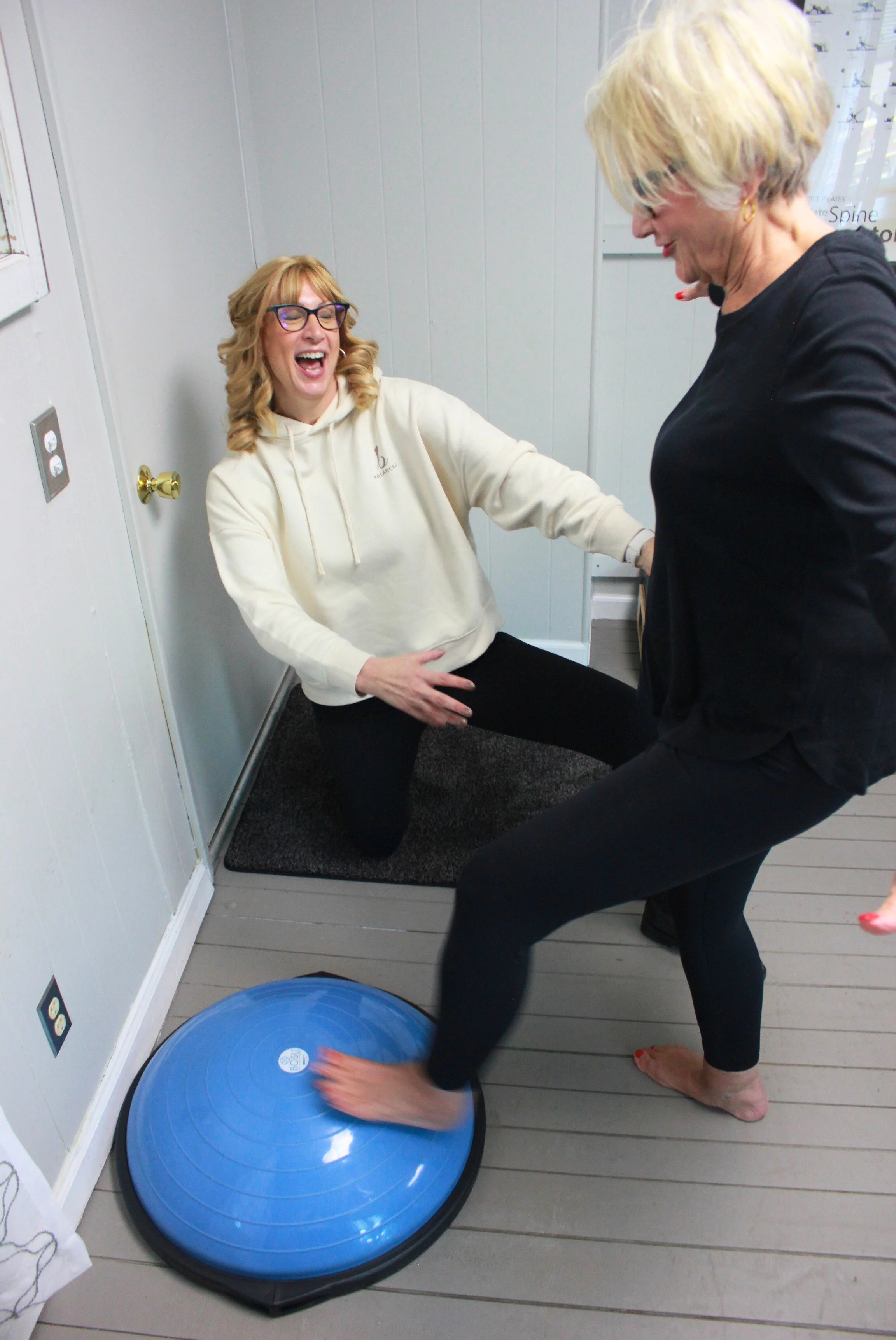 Two women are engaged in a playful activity in a brightly lit room. One woman with glasses and blonde hair is laughing and sitting on the floor, while the other woman, also blonde, is barefoot, standing, and moving a blue exercise ball with her foot.