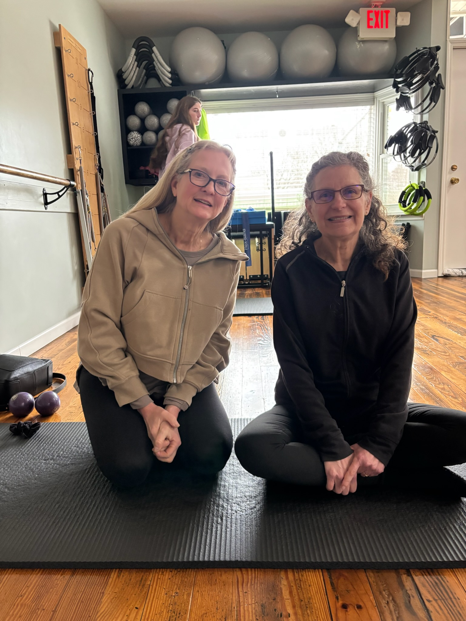 Two women sitting on Pilates mats at Balanced Pilates and Wellness Boutique in Midland, smiling at the camera with exercise equipment in the background, including stability balls, dumbbells, and resistance bands, with a young girl near the window.