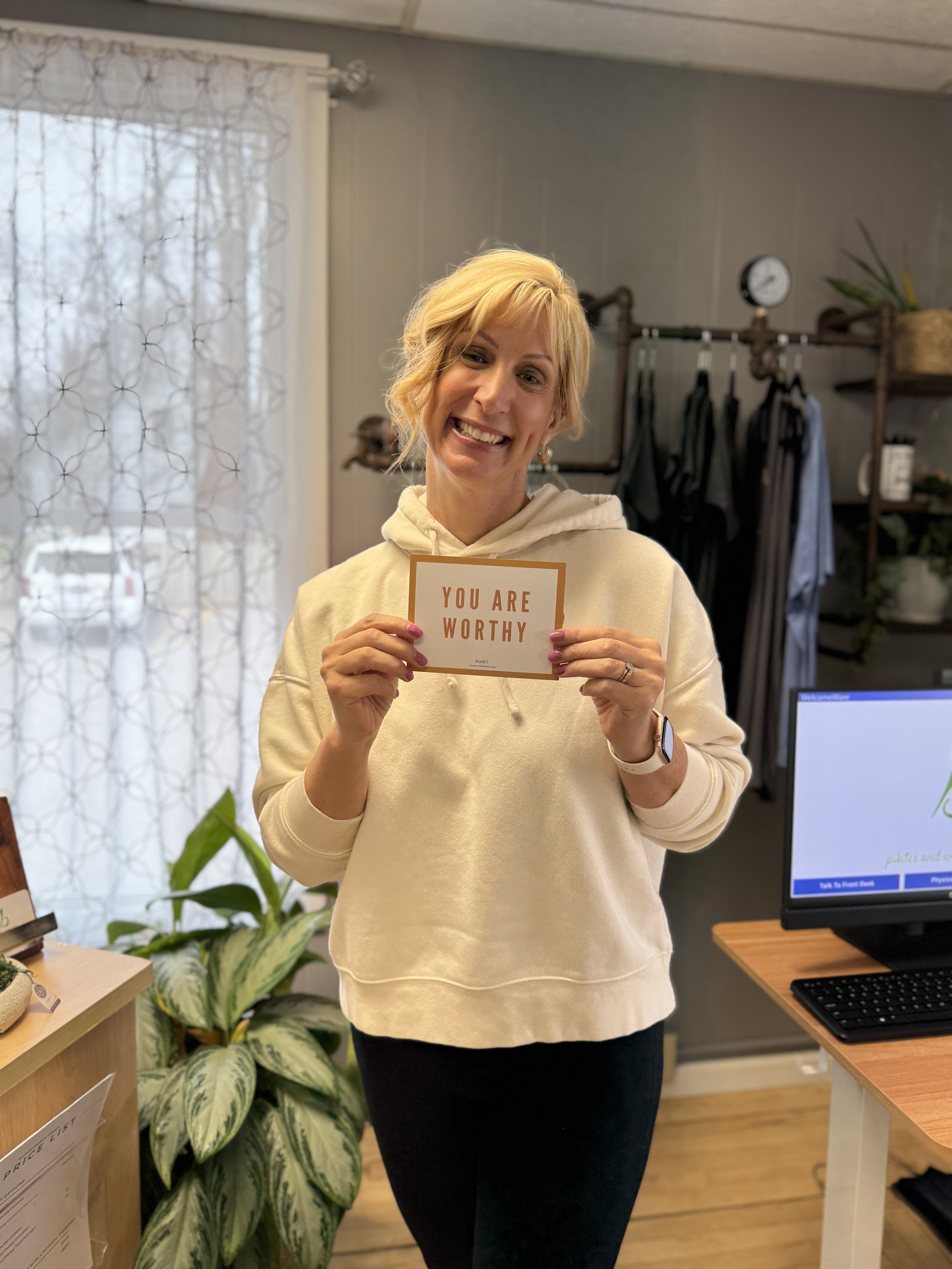 A woman with short blonde hair in a beige hoodie holding a card that reads "You Are Worthy" smiling at the camera in a cozy indoor setting at Balanced Pilates and Wellness Boutique in Midland, Michigan.
