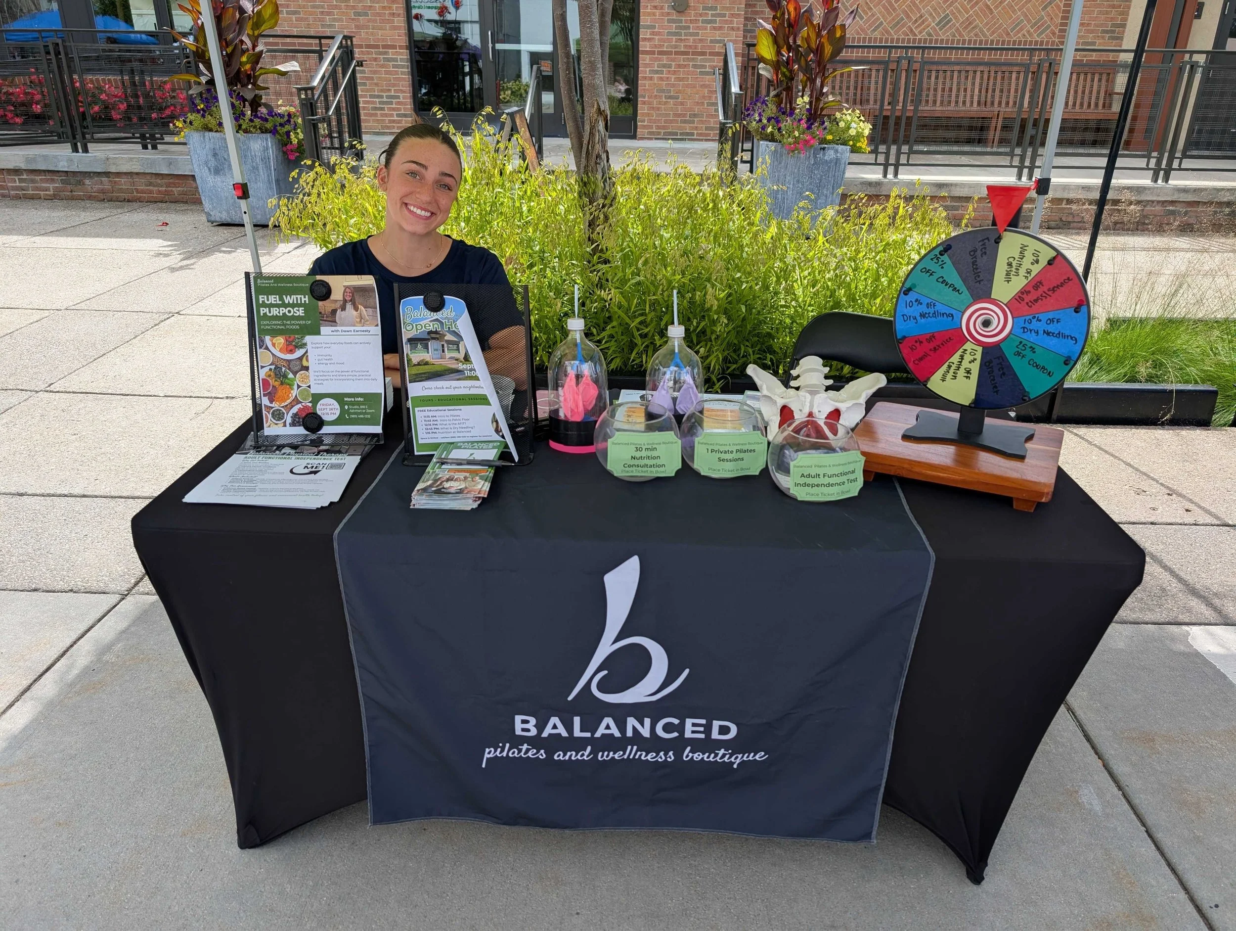 A woman sitting at a promotional booth with various informational materials and a prize wheel, outdoors in front of a brick building with plants and stairs.