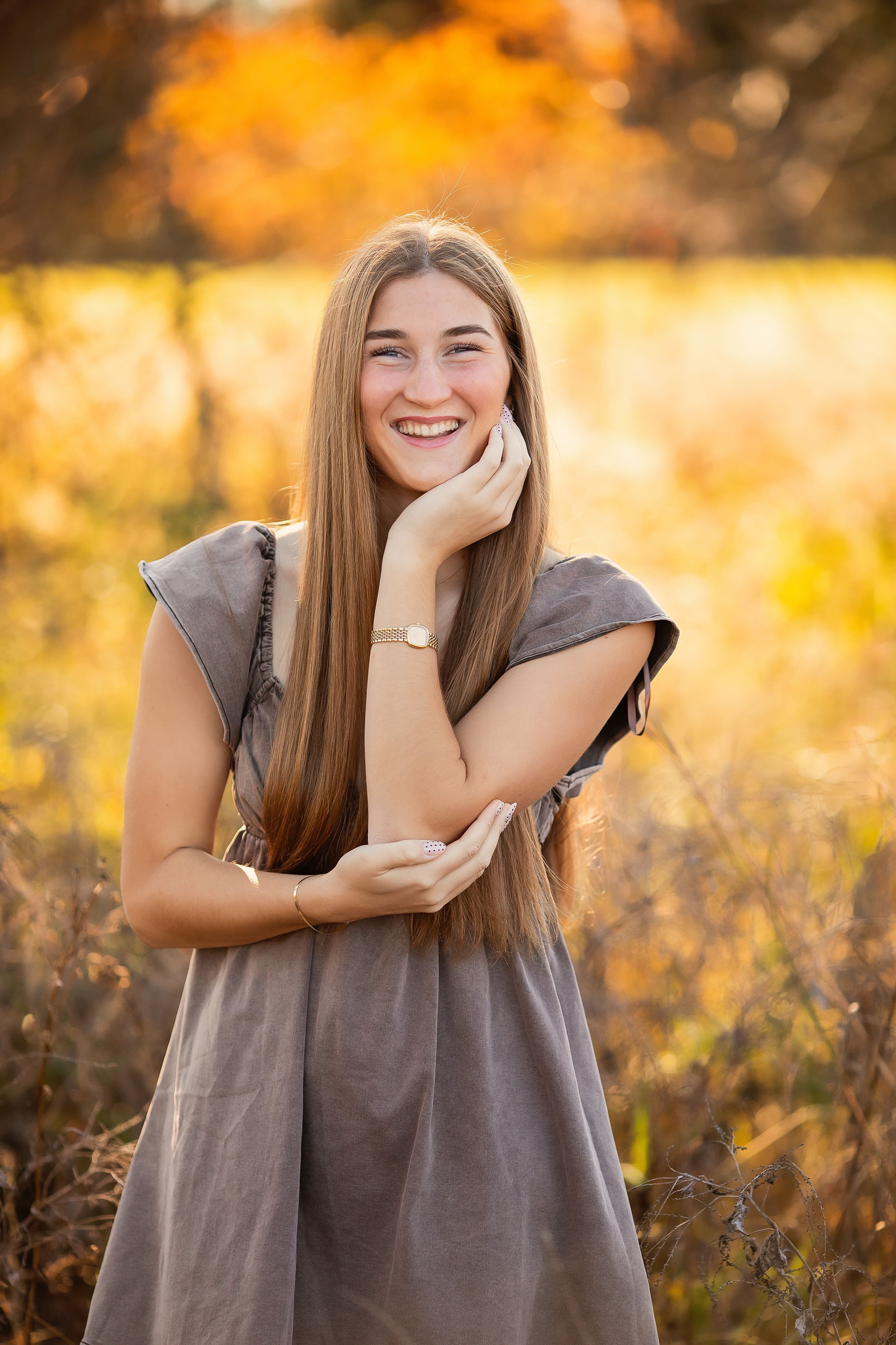 A young woman with long red hair, smiling, outdoors during autumn with yellow-orange trees and foliage in the background.