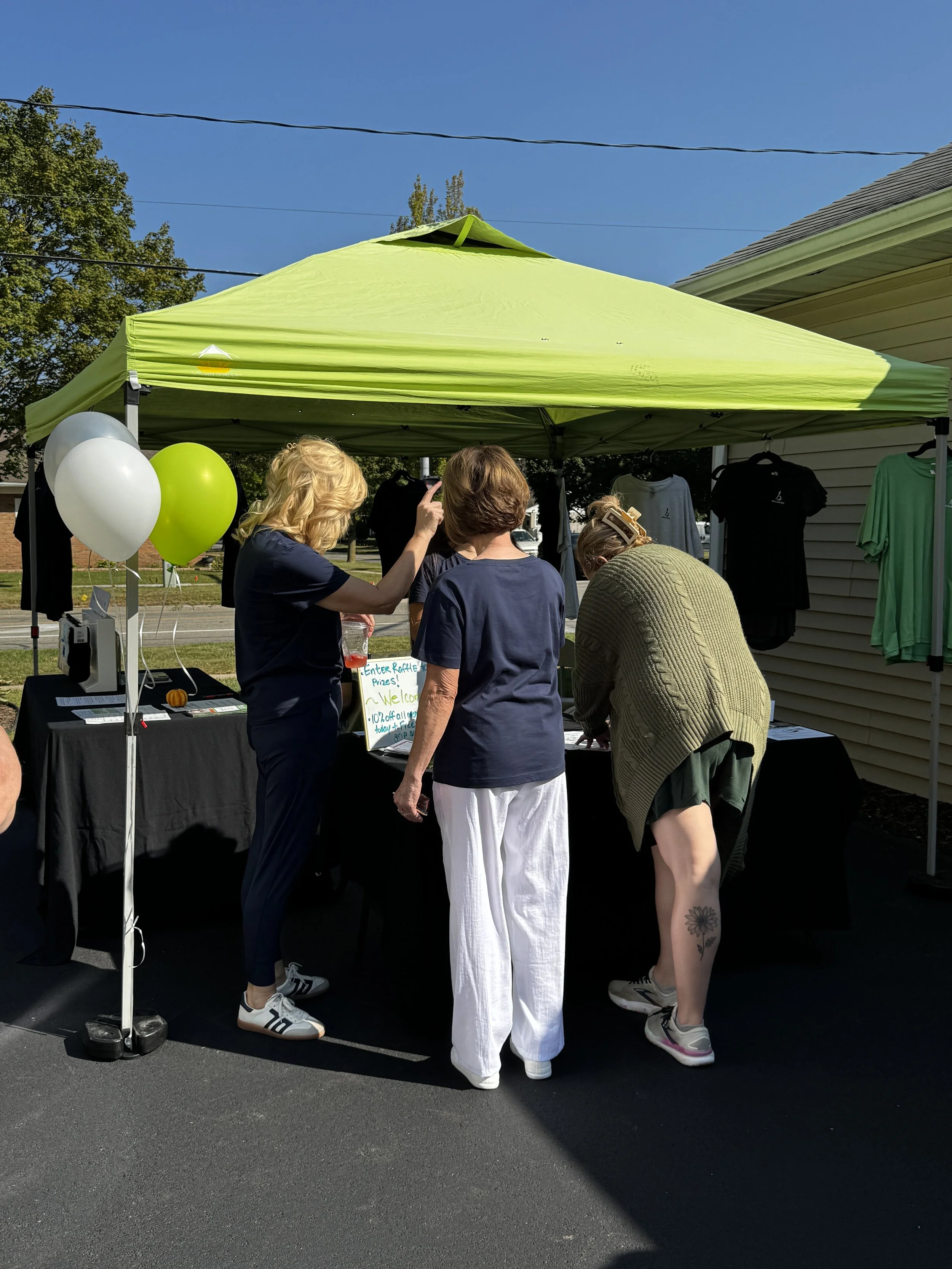 People gathered around a booth under a bright green canopy at Balanced Pilates and Wellness Boutique in Midland, Michigan, with balloons and clothing on display.