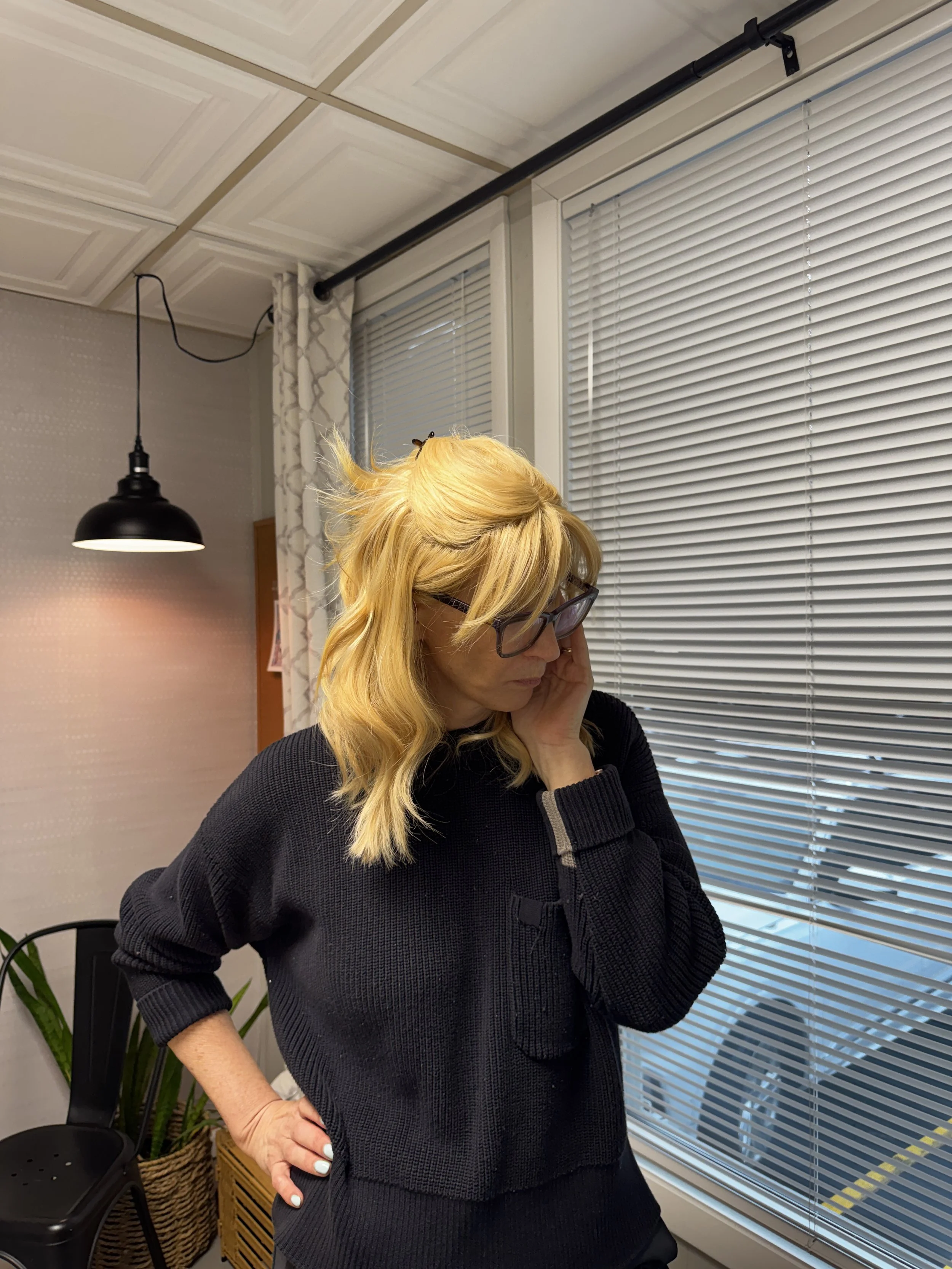 A woman with blonde hair and glasses, wearing a black sweater, standing indoors by a window with closed blinds, touching her face with her left hand.