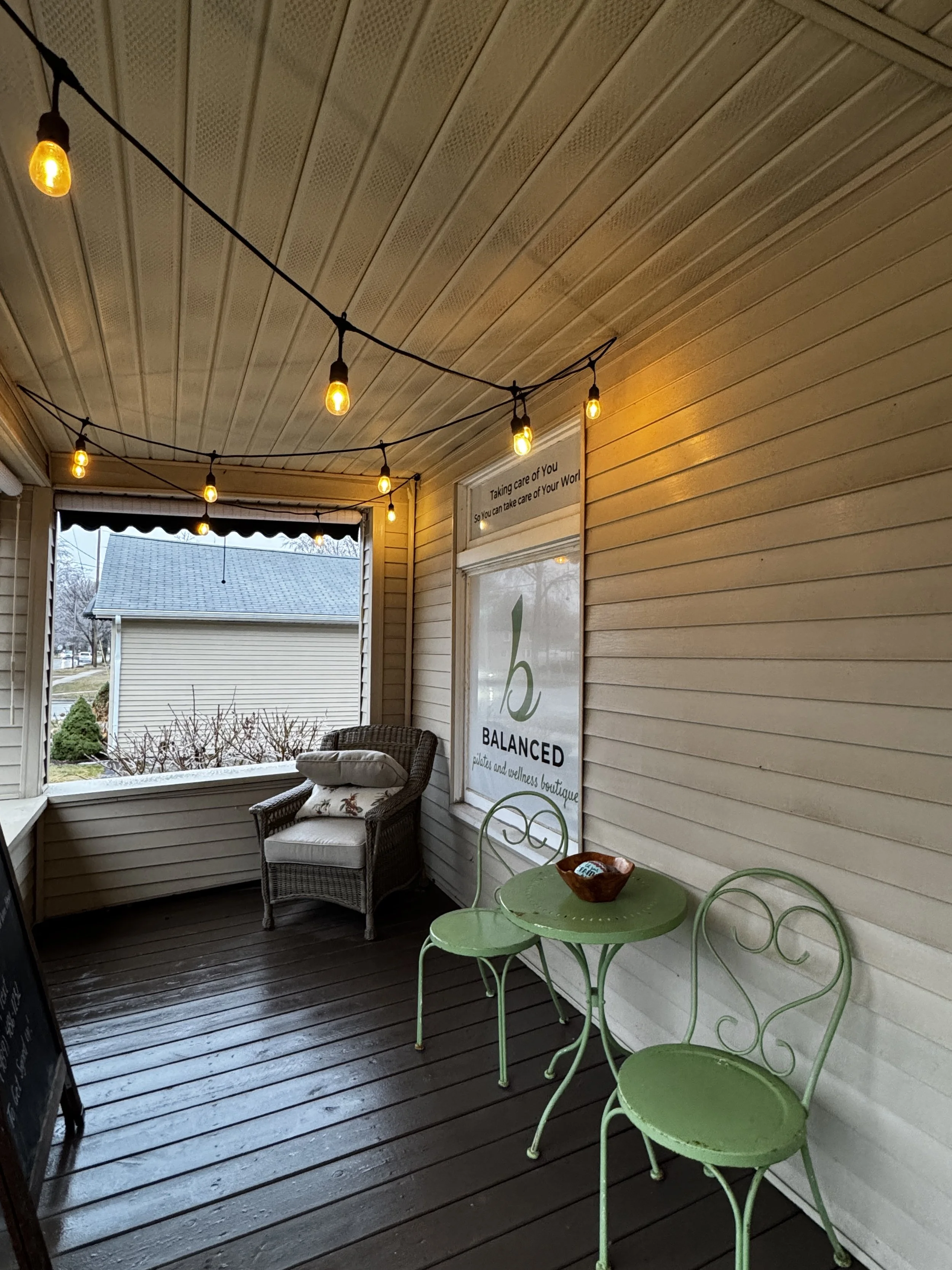 A porch with hanging string lights, a wicker armchair with pillows, a small green table and matching chairs, exterior walls with horizontal siding, and a large window with a sign that reads 'Taking care of you so you can take care of your world.'