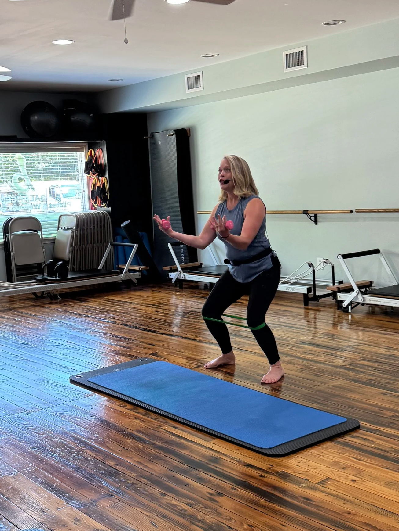 A woman instructs a STOTT Pilates class in the Balanced studio, using a resistance band around her thighs while performing a squat on a blue yoga mat.