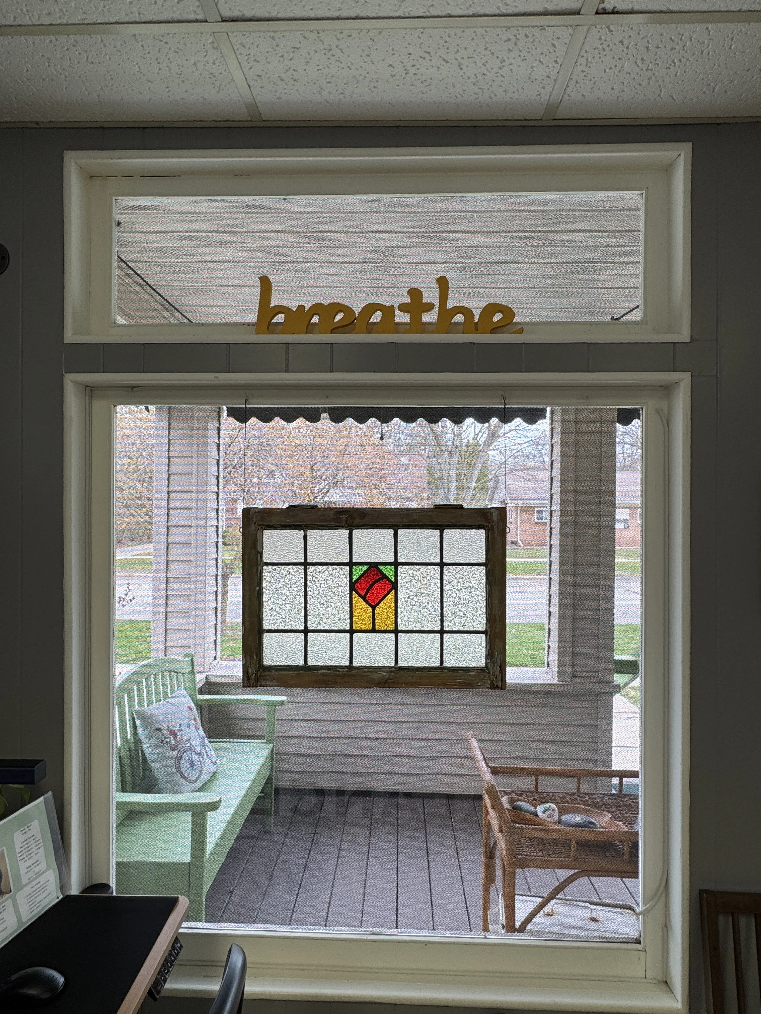 View through a window showing a porch or enclosed sunroom with chairs, a decorative pillow, and a stained glass window with a red, yellow, and green geometric design.