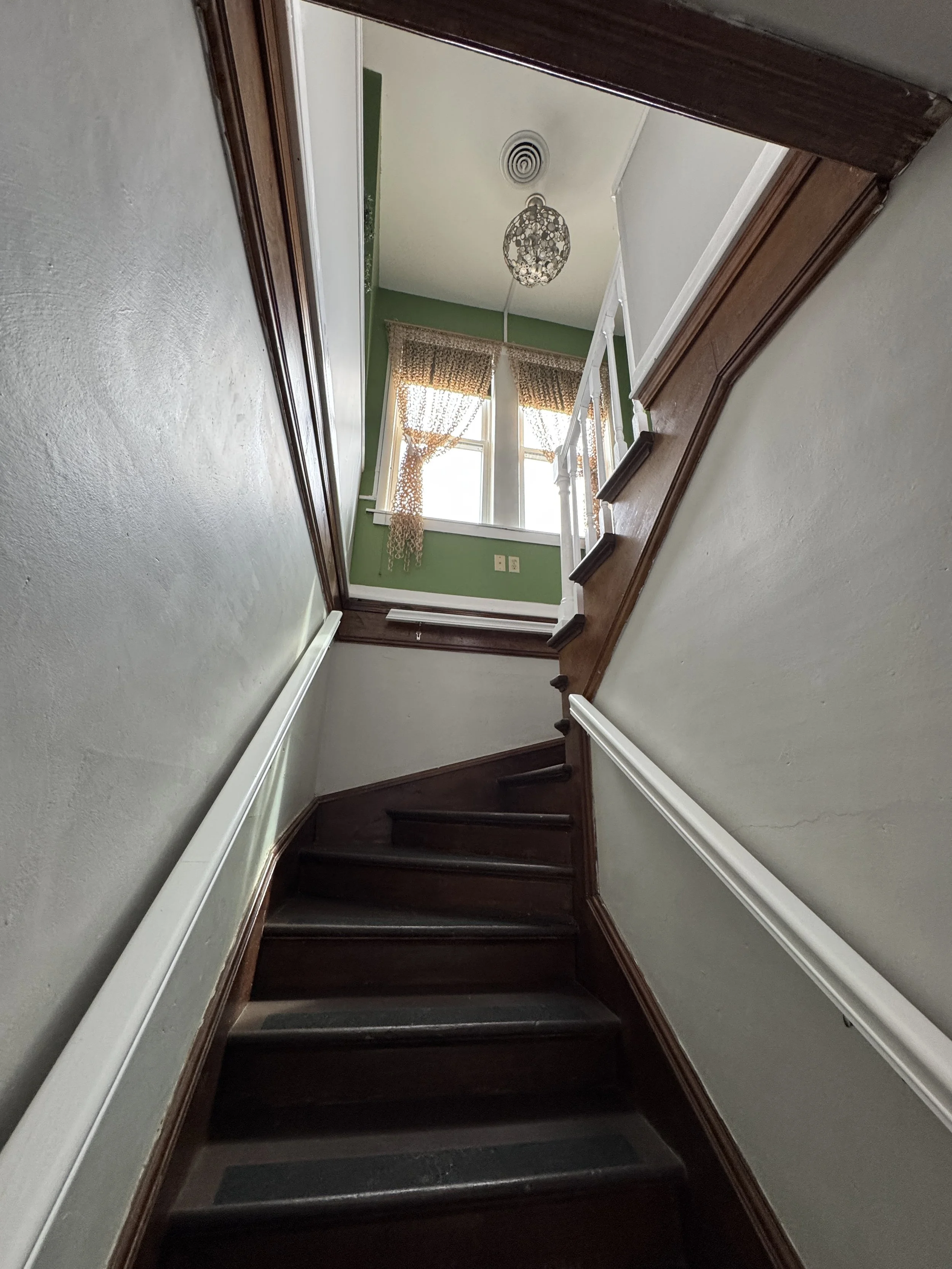 Interior view of a staircase from the bottom, showing dark wooden stairs, white walls with a chair rail, and a green room at the top with windows and curtains, with a ceiling light fixture.