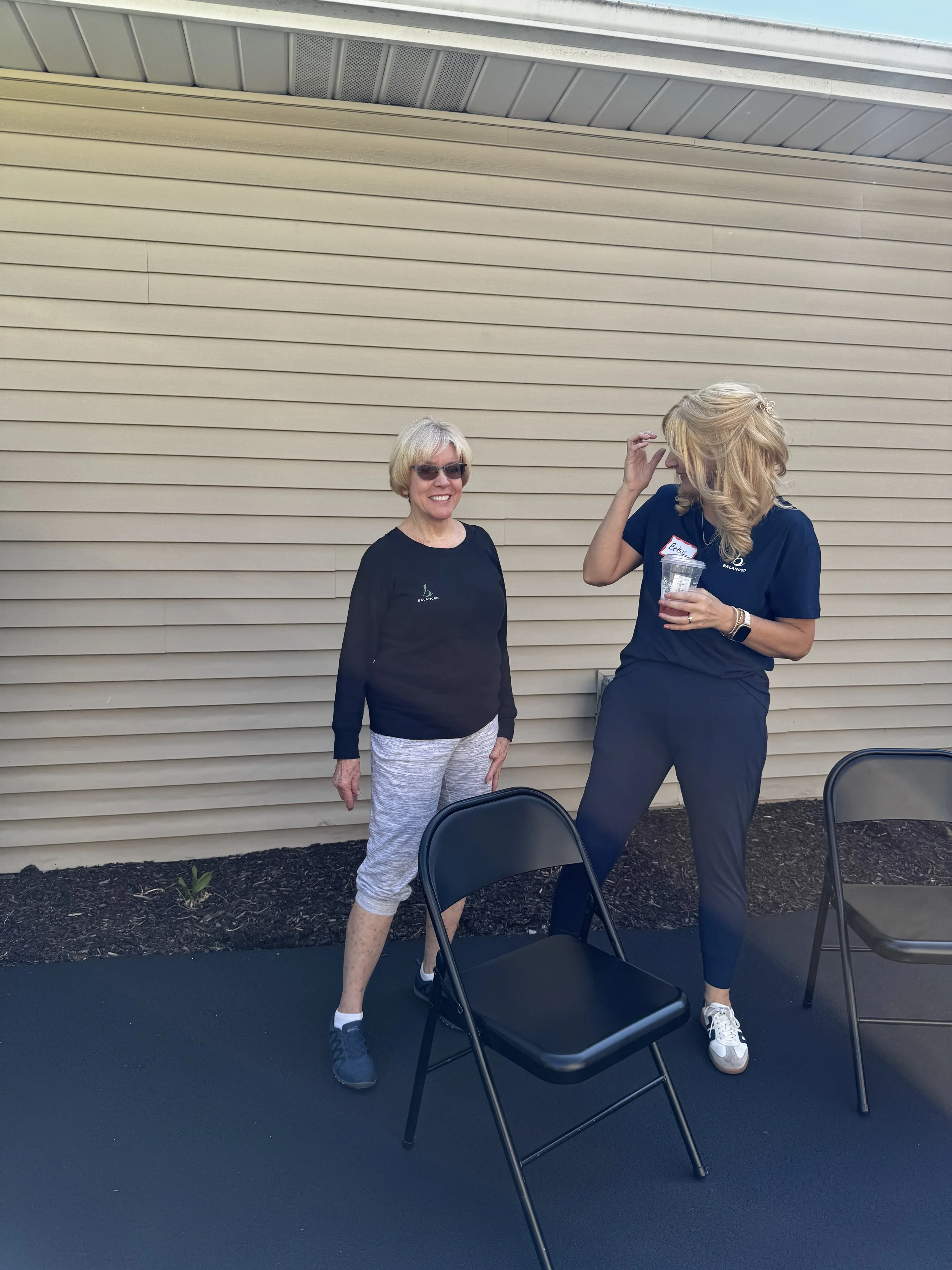 Two women standing outdoors against a beige horizontal siding wall, with black metal chairs in front of them. The woman on the left has short blonde hair, glasses, a black shirt, gray shorts, and black shoes. The woman on the right has shoulder-lengt