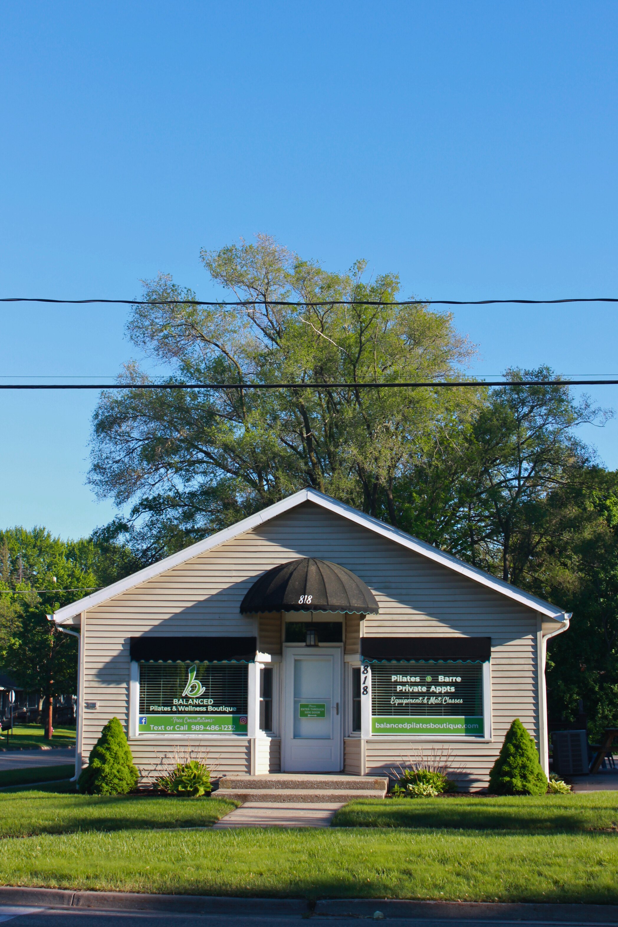 Small beige building with a black awning over the entrance, green signs on the windows indicating Pilates and wellness services, surrounded by a well-maintained lawn and bushes, with trees and a clear blue sky behind.