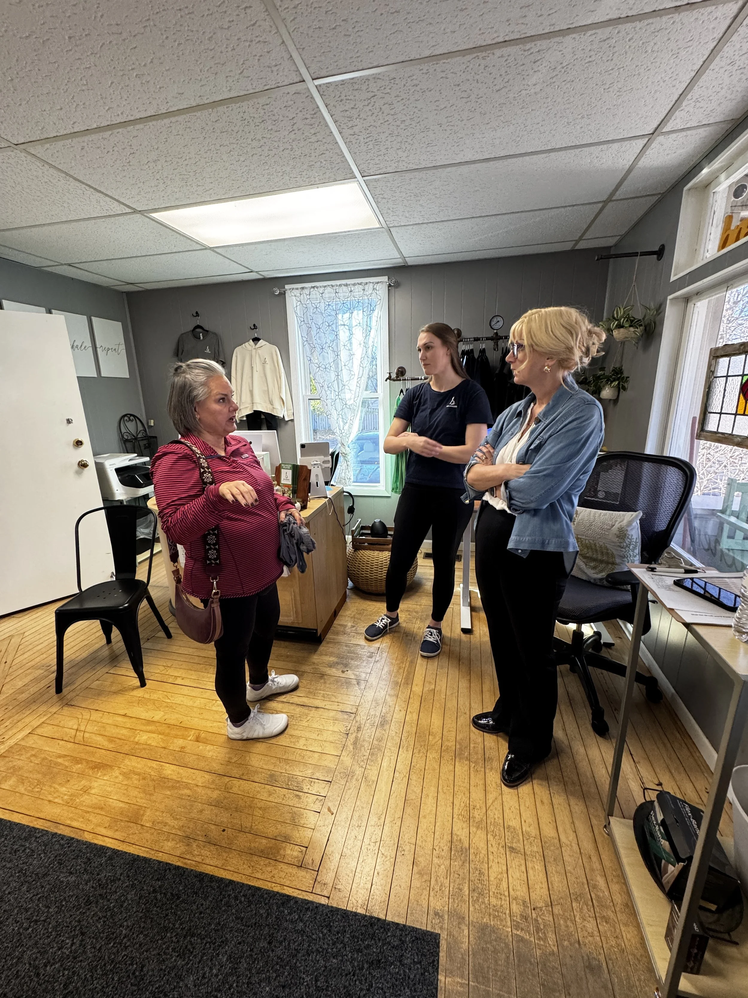 Three women having a conversation at Balanced Pilates and Wellness Boutique in Midland, Michigan. One woman in a striped shirt is speaking, while the other two women are listening, standing with arms crossed.