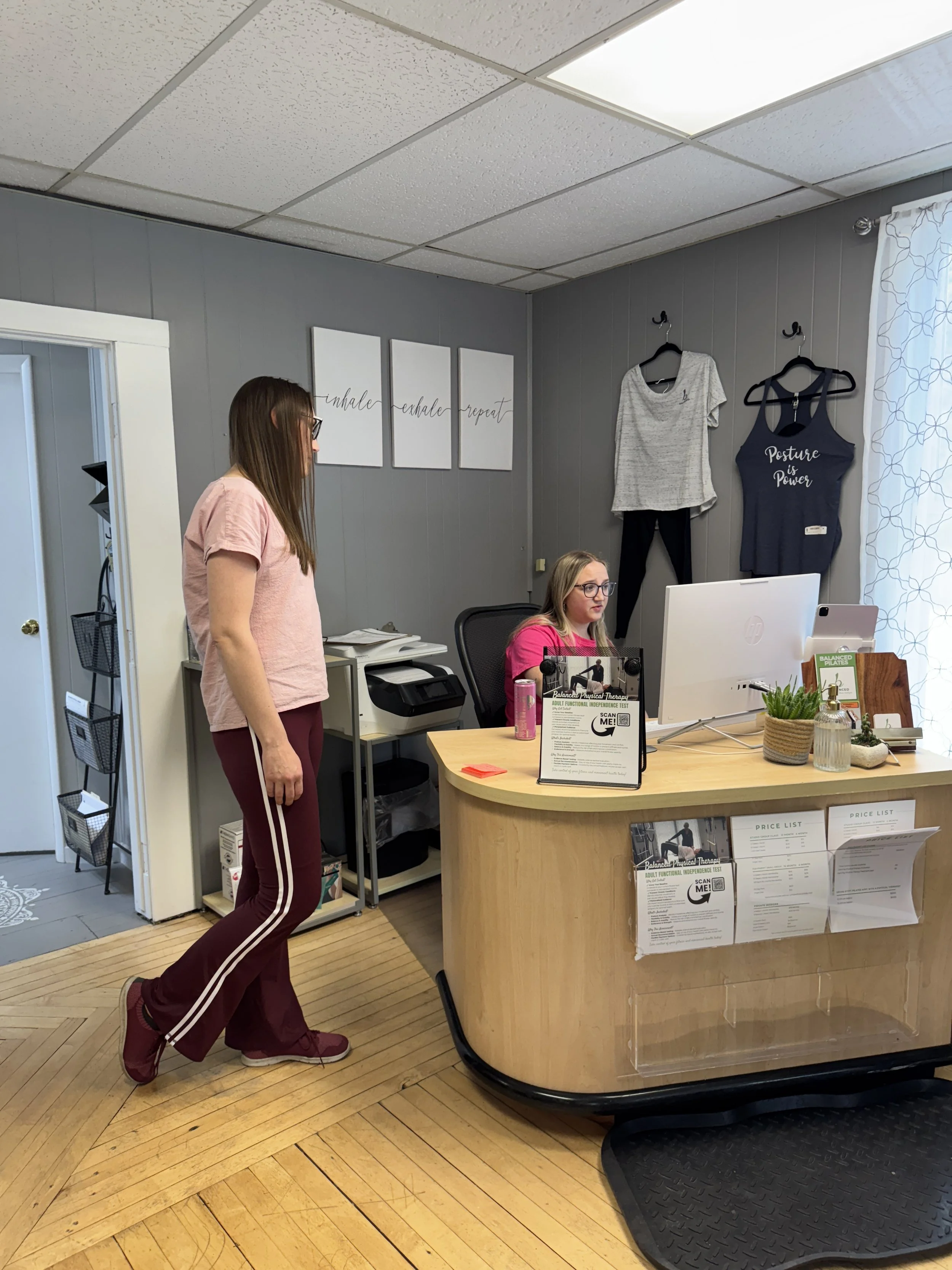 Two women in an office, one standing and one sitting at a desk, with a computer and various office supplies. Behind them, the wall has three framed motivational posters, and two pieces of clothing hanging on hooks. The room has gray walls, a wood flo