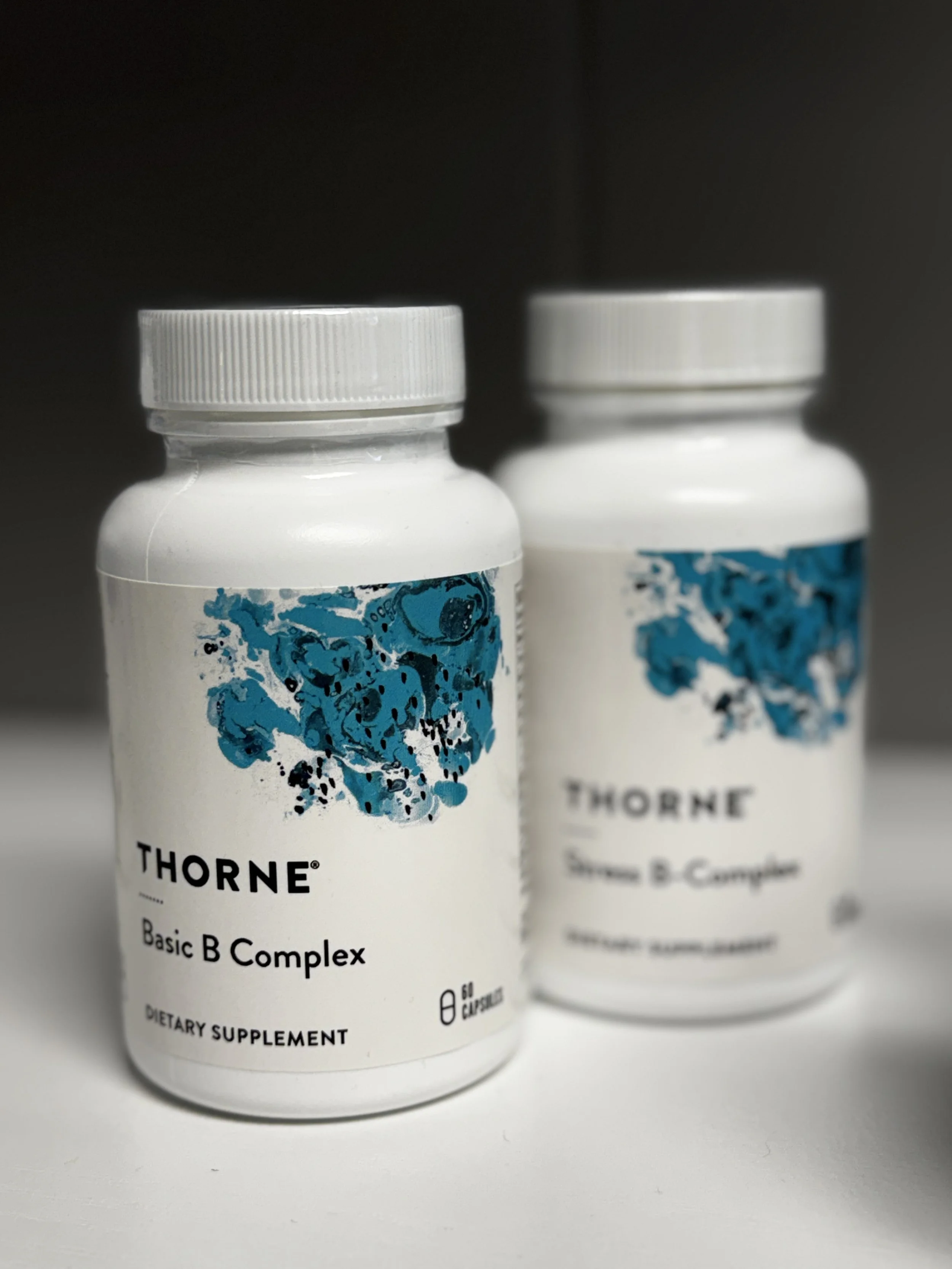 Two white bottles of Thorne Basic B Complex dietary supplements with blue abstract designs on the labels, placed on a white surface against a dark background.