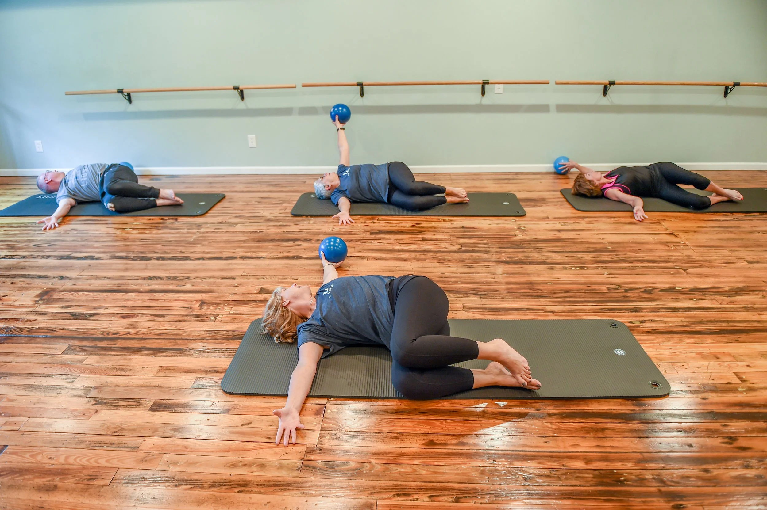 People practicing STOTT Pilates on mats in a studio with wooden floors and wall bars, some holding blue exercise balls at Balanced Pilates and Wellness Boutique in Midland, Michigan