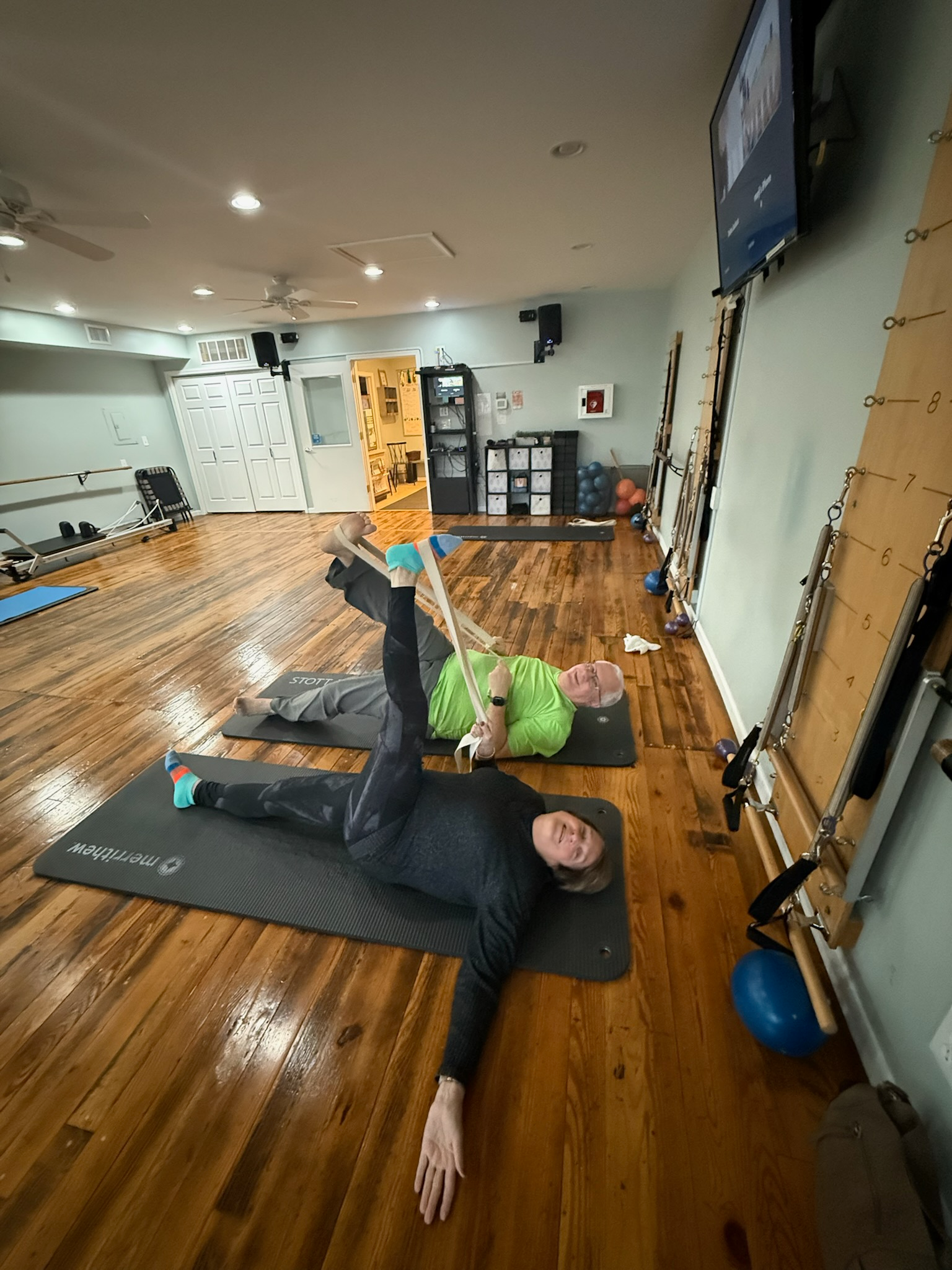 Indoor Pilates studio with two people performing stretching exercises on yoga mats. The room has a wooden floor, exercise equipment, and a large screen mounted on the wall.