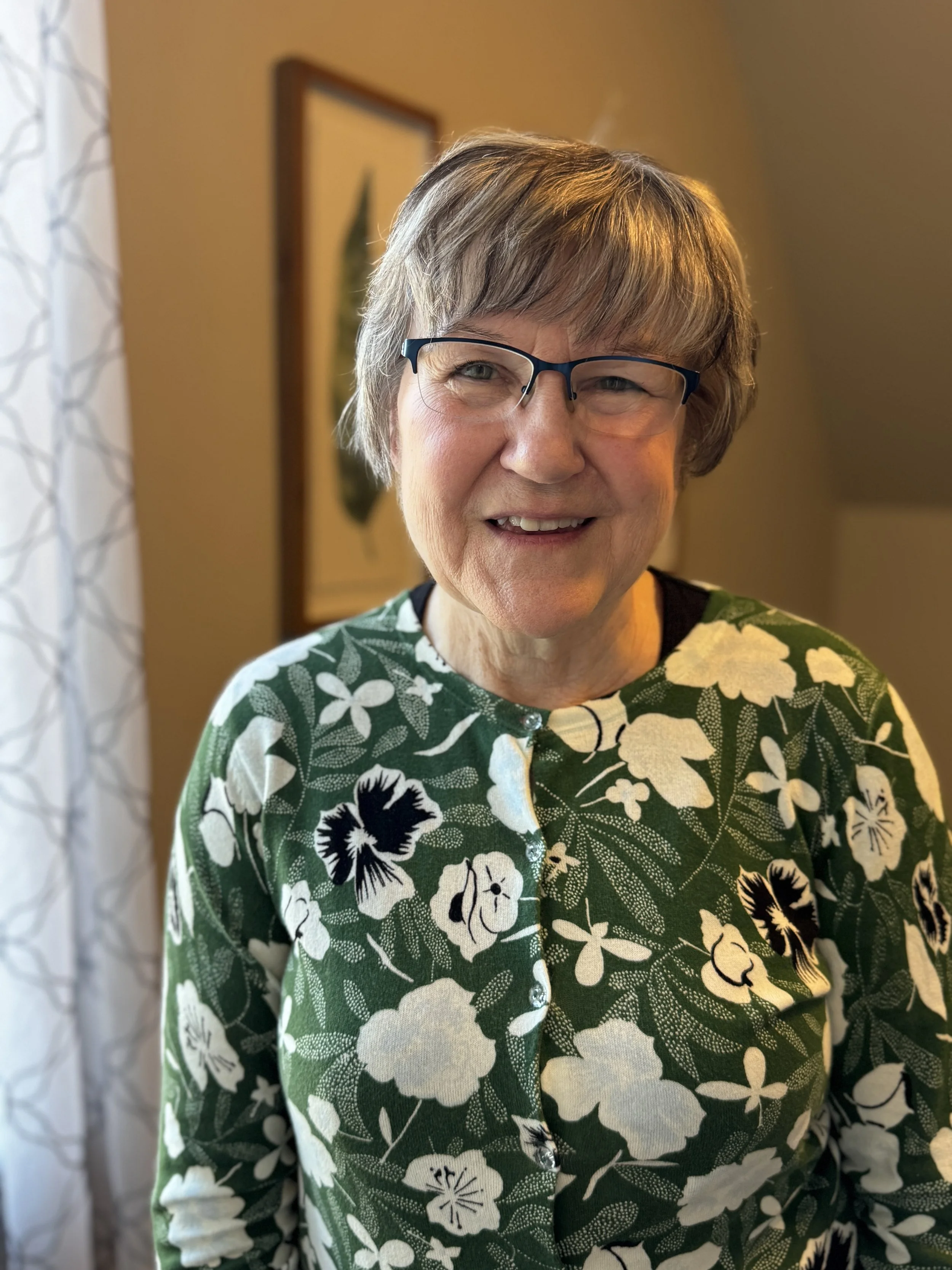 Close-up of an older woman with short gray hair, wearing glasses and a green floral-patterned shirt, smiling indoors.