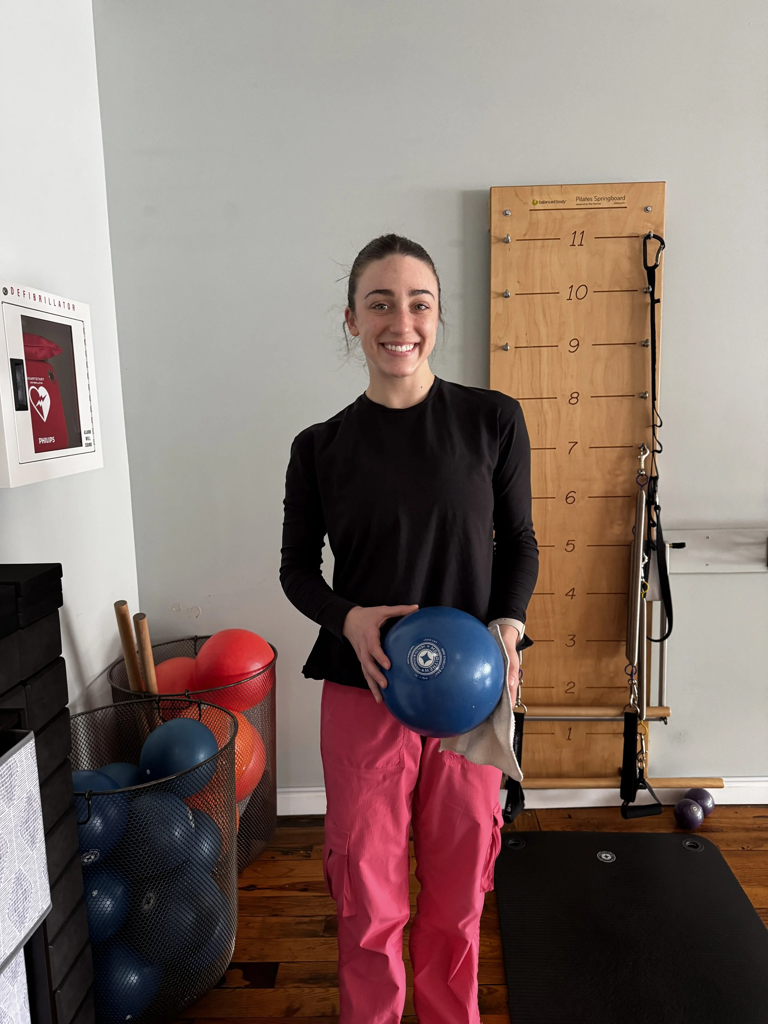 A young woman with brown hair pulled back, smiling, holding a blue exercise ball in a gym or workout space. She is wearing a black long-sleeved shirt and pink pants. Behind her is a wooden height measurement wall, resistance bands, and fitness equipm