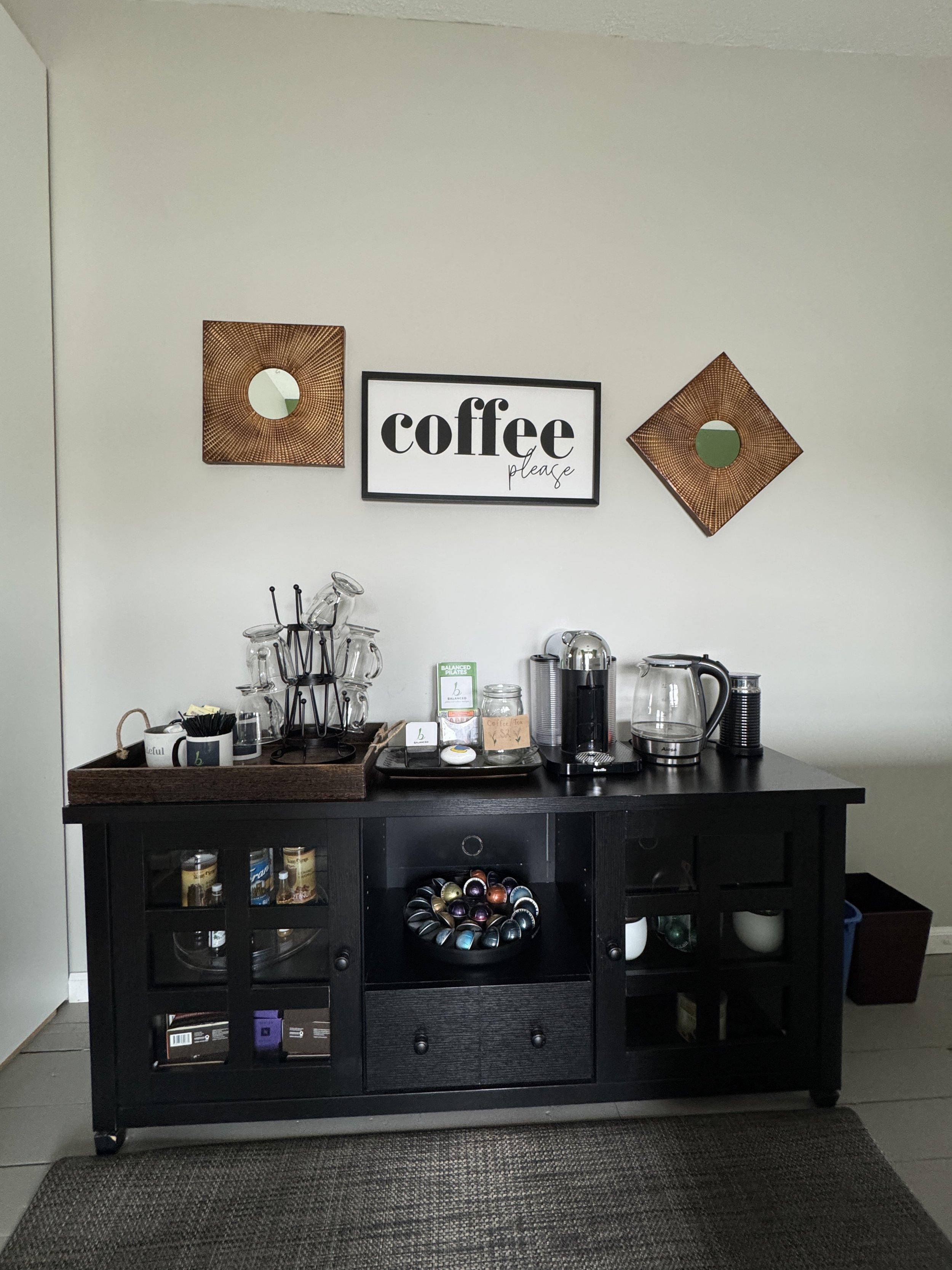 A black wooden cabinet with glass doors holding coffee supplies, and a coffee station on top including a coffee machine, kettle, and various cups, with a decorative framed sign that says 'coffee please' above it. Two square copper-colored mirrors wit