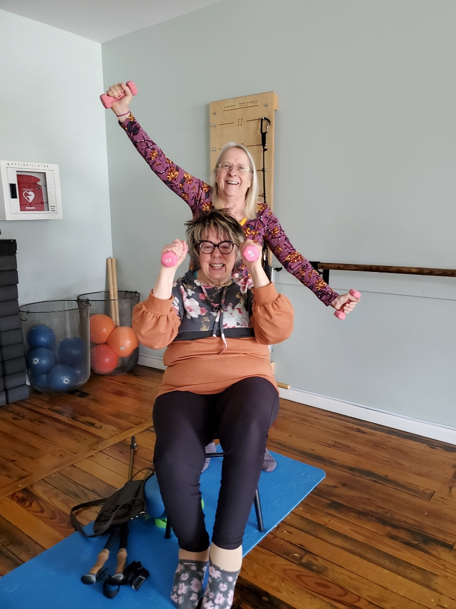 Two women in workout attire, one sitting on a chair and the other standing behind, holding dumbbells and lifting a pink dumbbell with one arm. They are smiling and holding Pilates props at Balanced Pilates and Wellness Boutique in Midland, Michigan.