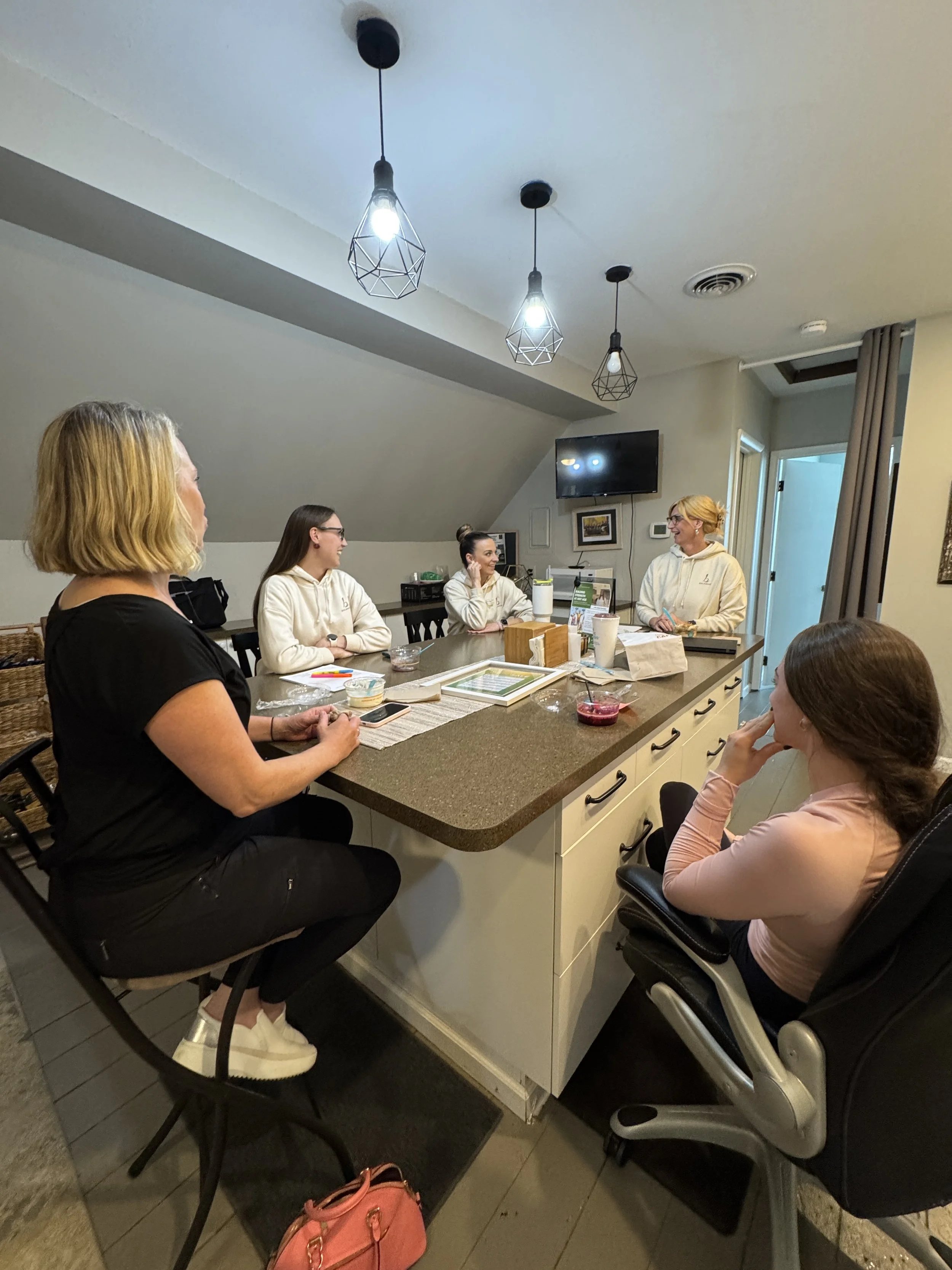 A group of six women sitting around a kitchen island, engaged in conversation. The women are of various ages, some with glasses, and are dressed casually. There is a pink purse below the island and a few dishes and cups on the counter. The kitchen ha