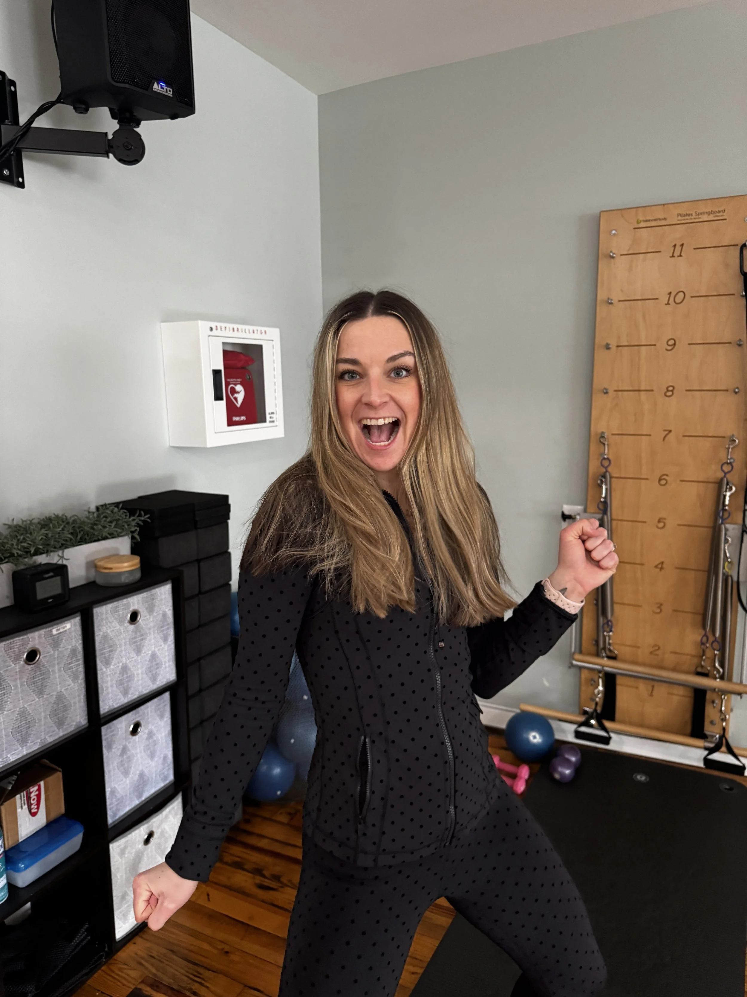 A woman with long blonde hair in workout clothes is smiling and in a squatting position in a home gym, surrounded by fitness equipment such as medicine balls, resistance bands, and a wall-mounted wooden climbing frame.