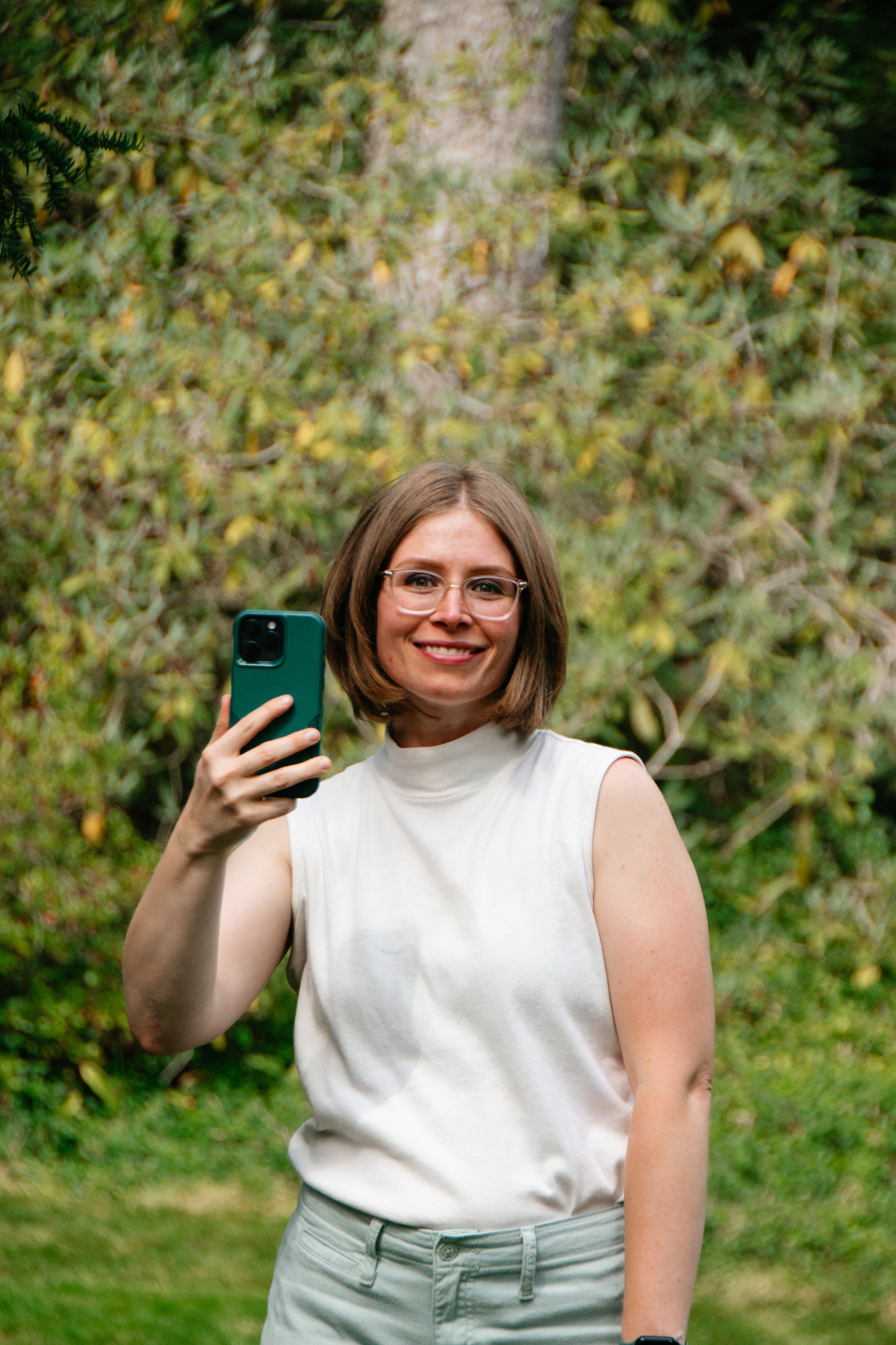 Kathleen Haney, marketing specialist, with glasses and a short bob haircut takes a selfie outdoors, smiling, with a wooded background.