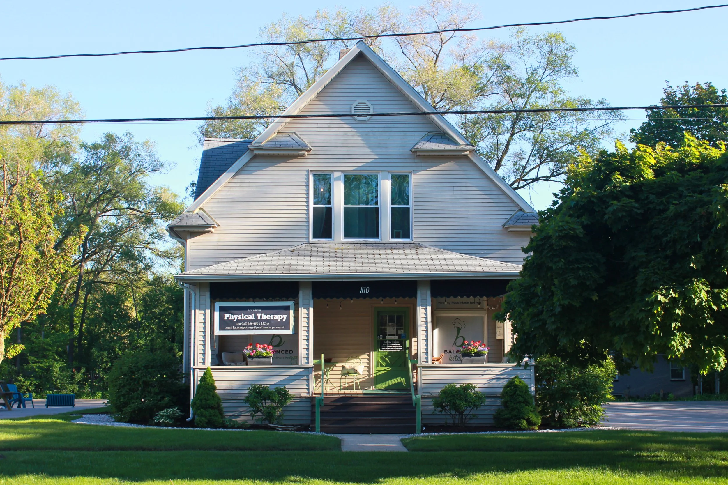 A two-story gray house with a small porch, surrounded by trees and greenery, with a sign for a physical therapy business on the porch at Balanced Pilates and Wellness Boutique in Midland, Michigan