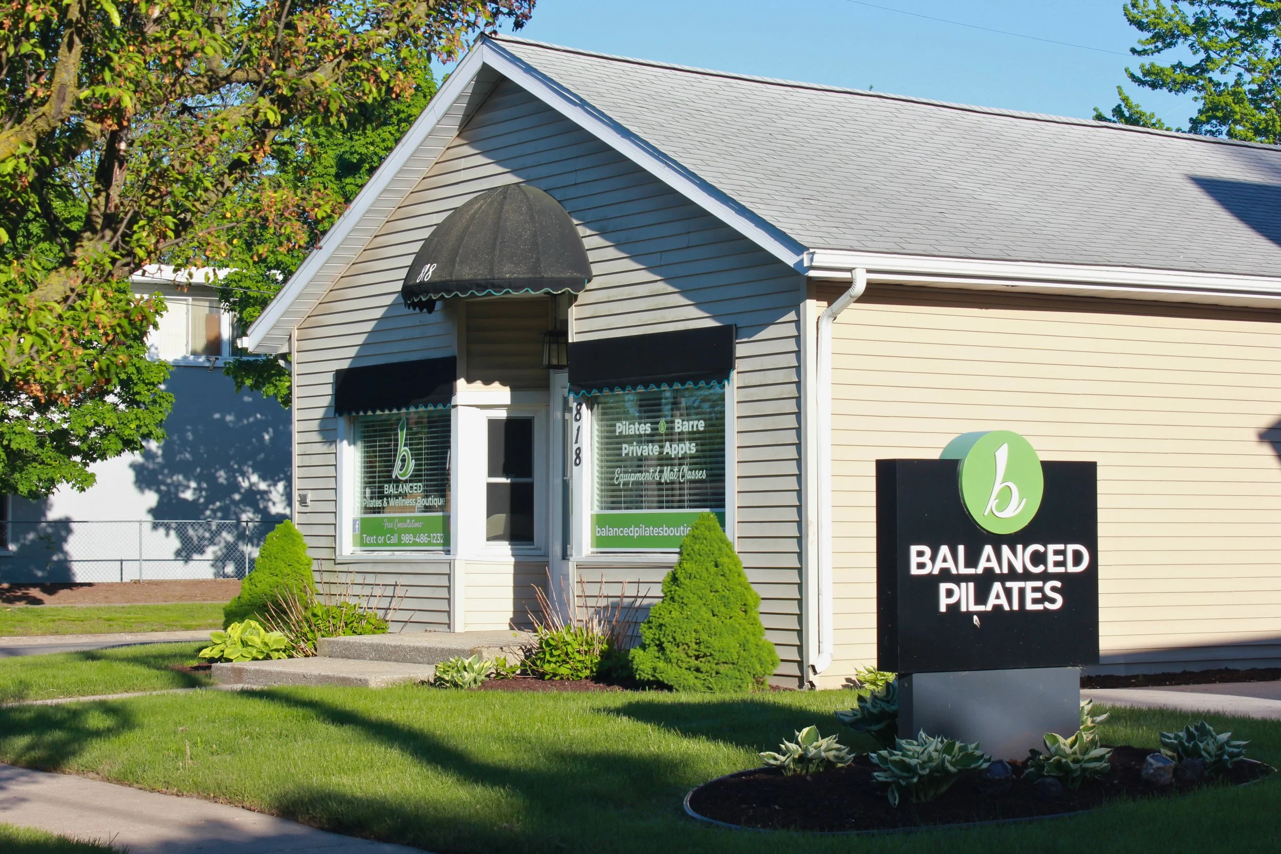 Exterior of a small business called Balanced Pilates, with a large window displaying the services offered, a hanging black awning over the door, and a sign in front advertising Pilates classes and private appointments. The building has beige siding, 
