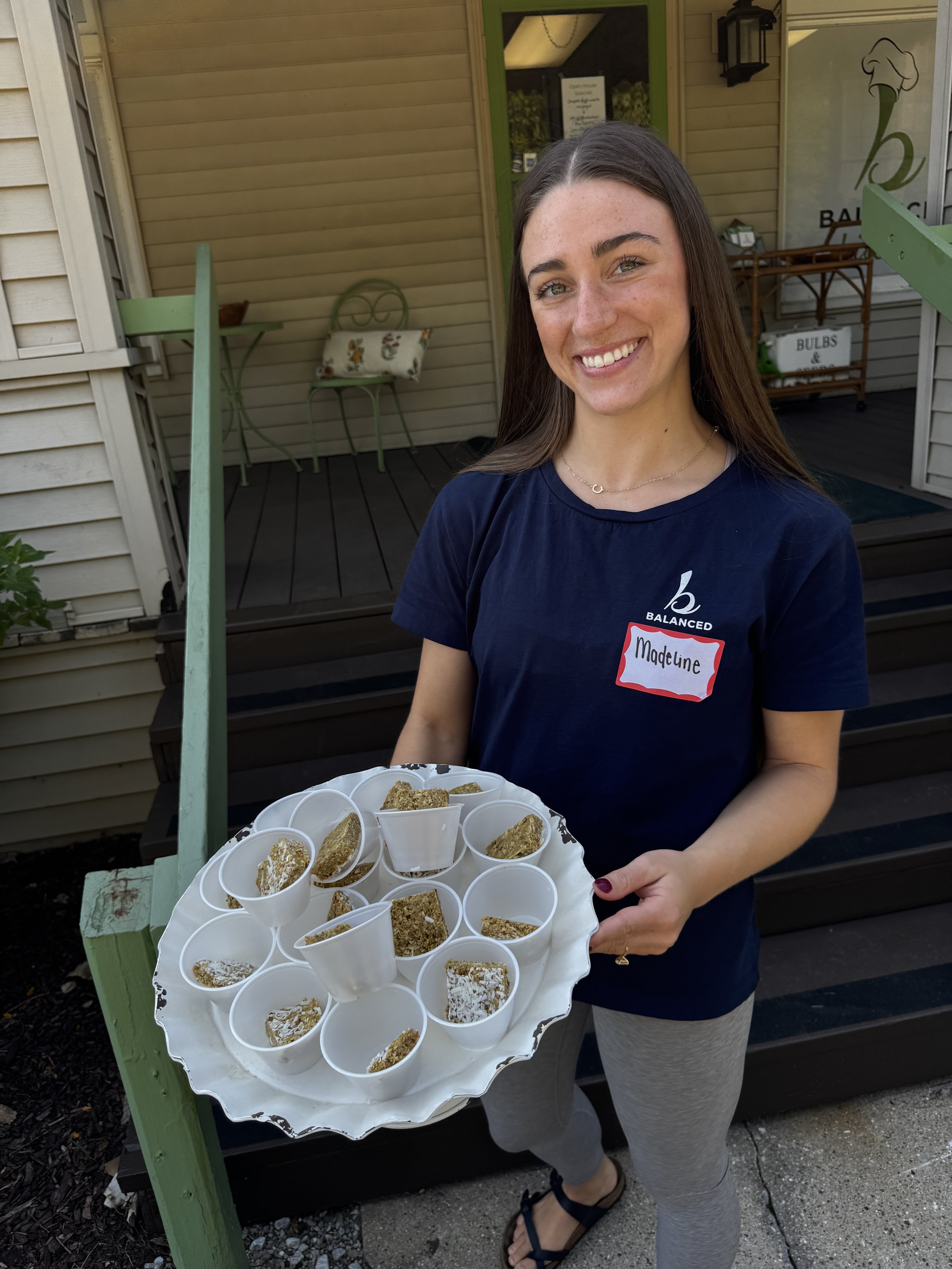 A woman holding a tray of small cups filled with baked goods, standing outside near a building with beige siding and a green door. The woman is smiling and wearing a navy T-shirt with a name tag that says "Madeline."